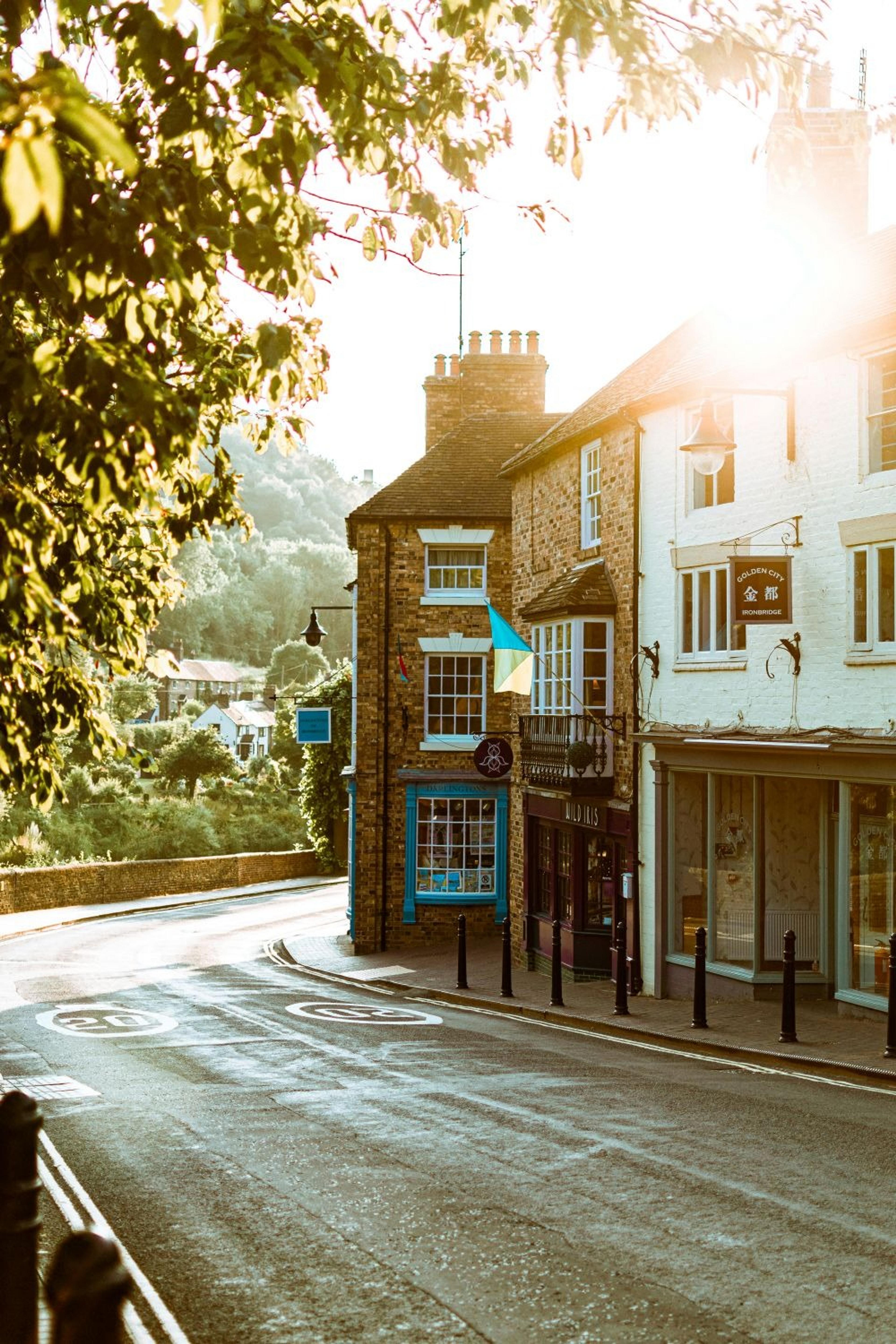 A quaint street bathed in warm sunlight in Ironbridge, Shropshire, with brick buildings and lush greenery in the background.