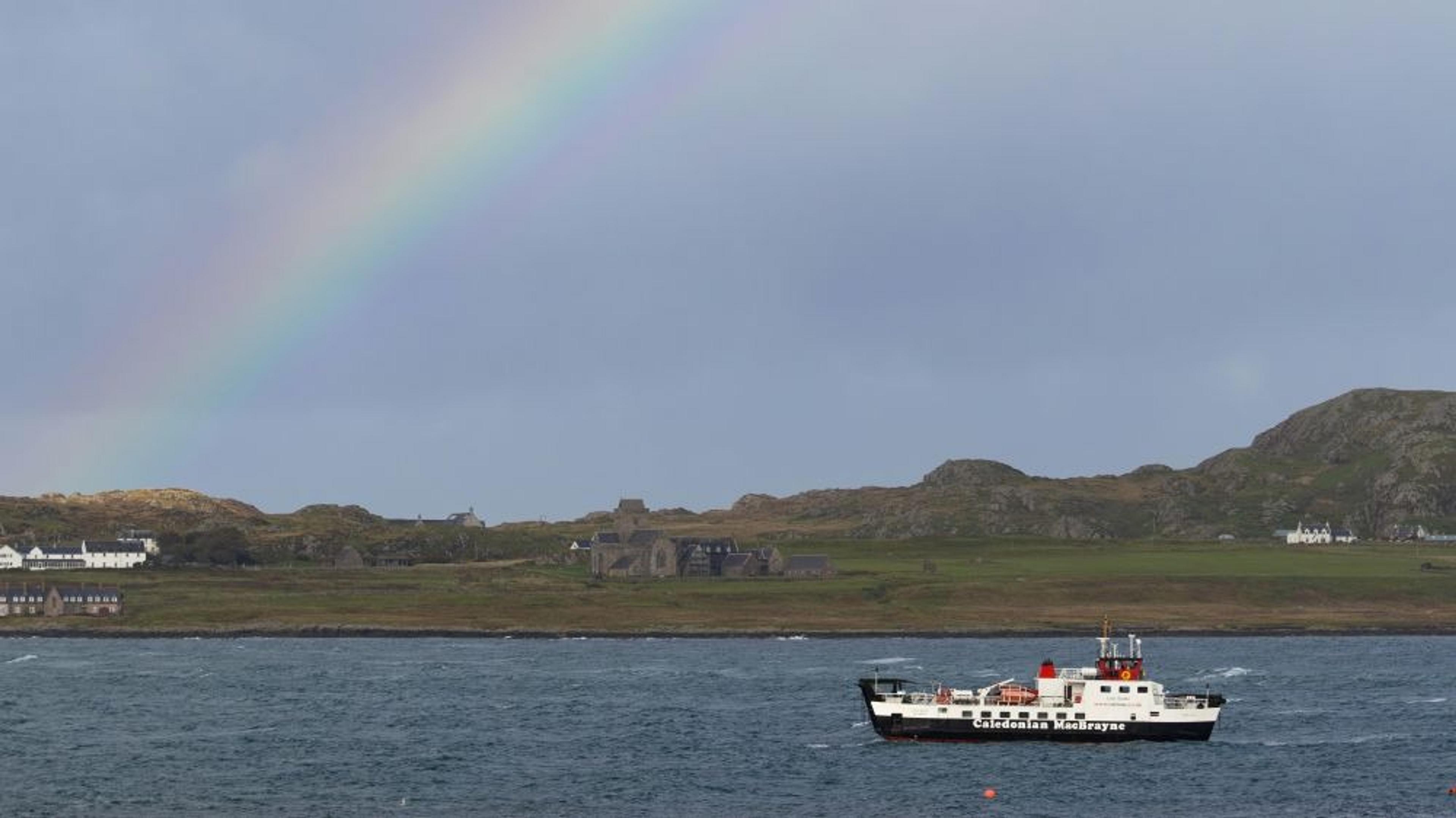 A ferry sails near the coast of Iona, Scotland, beneath a vibrant rainbow.