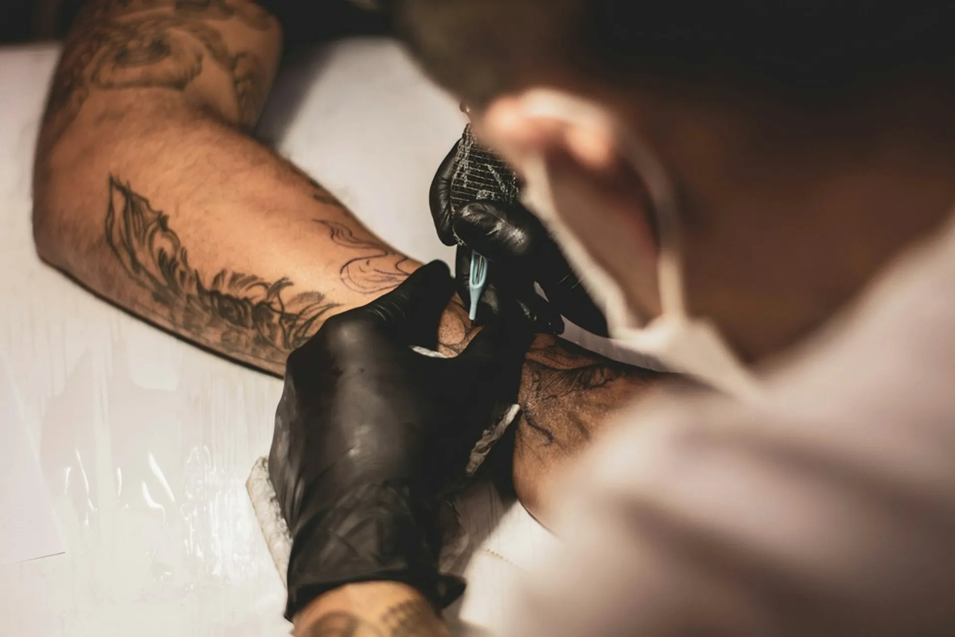 A tattoo artist wearing gloves and a mask carefully inks a client's arm in a studio.