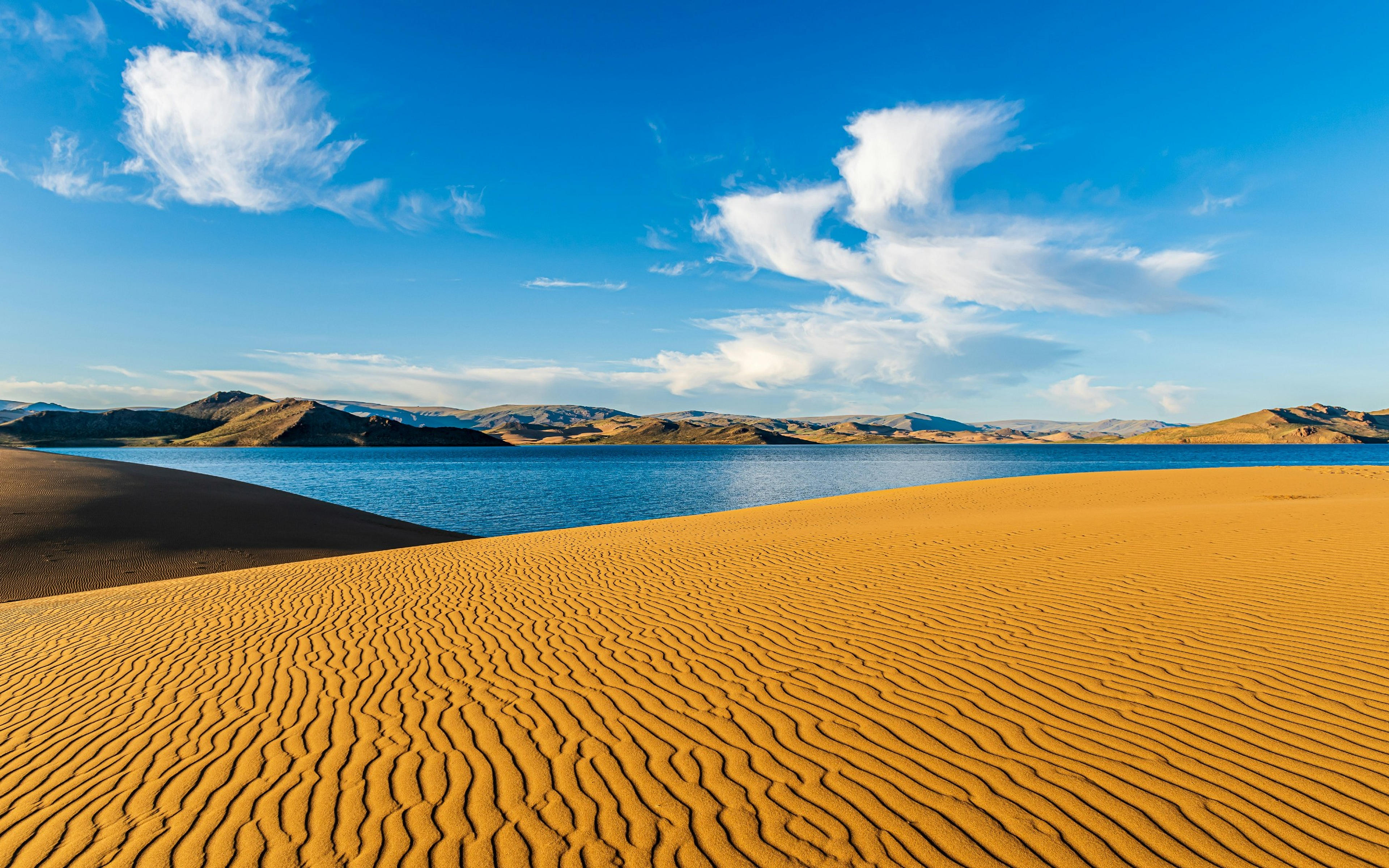 Golden sand dunes with intricate patterns overlook the serene waters of Khyargas Lake in Mongolia, framed by distant rolling hills.