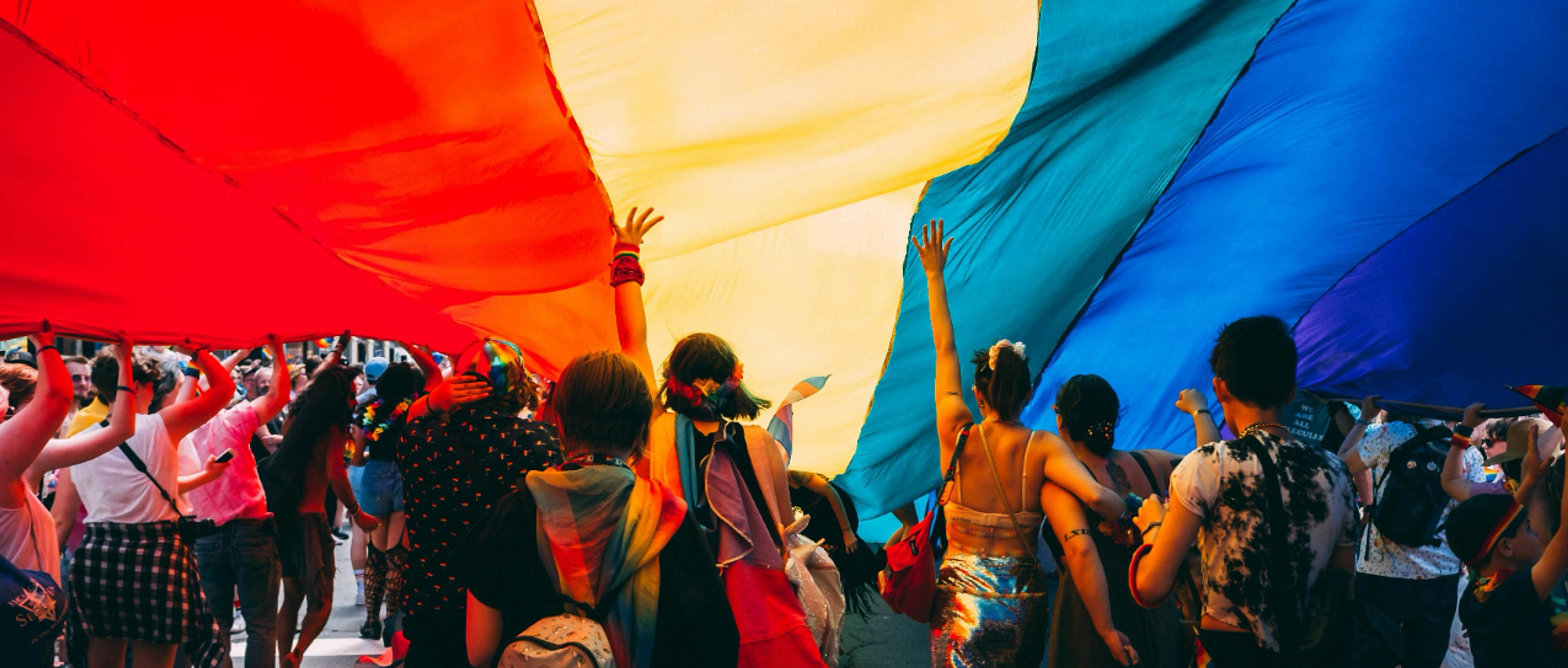 A vibrant group of people celebrates under a large rainbow flag during a Pride Parade.
