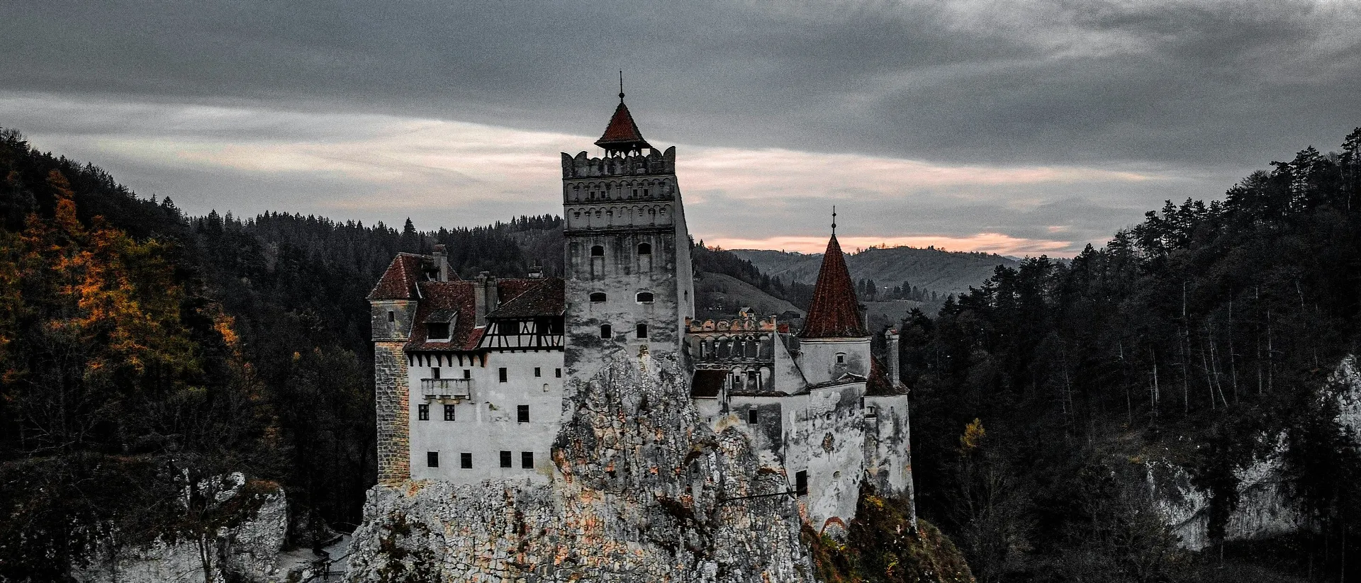 Bran Castle in Romania emerges dramatically from the surrounding dark forest under a moody sky.