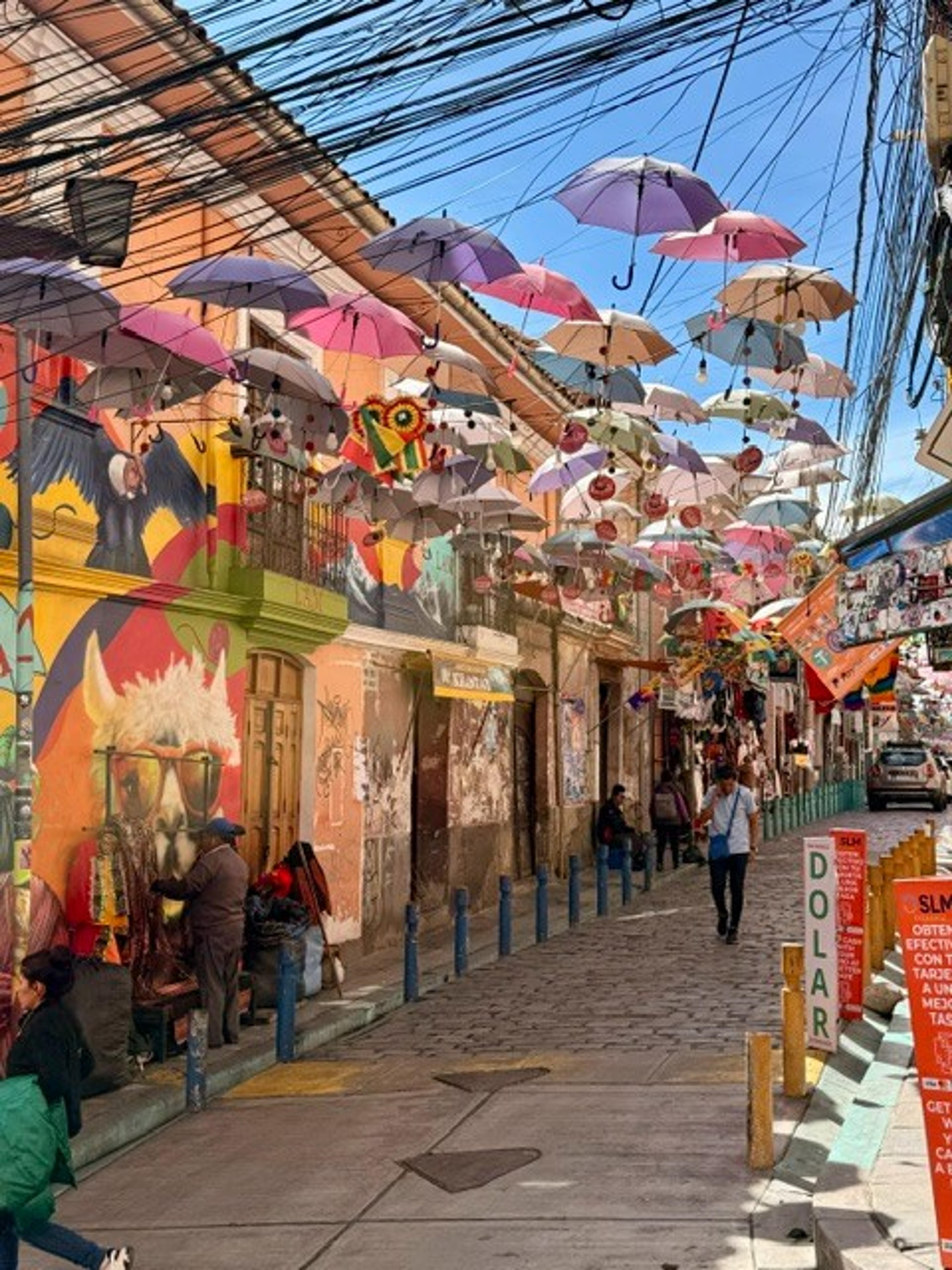 A vibrant street in La Paz, Bolivia is adorned with colorful murals and umbrellas suspended overhead.