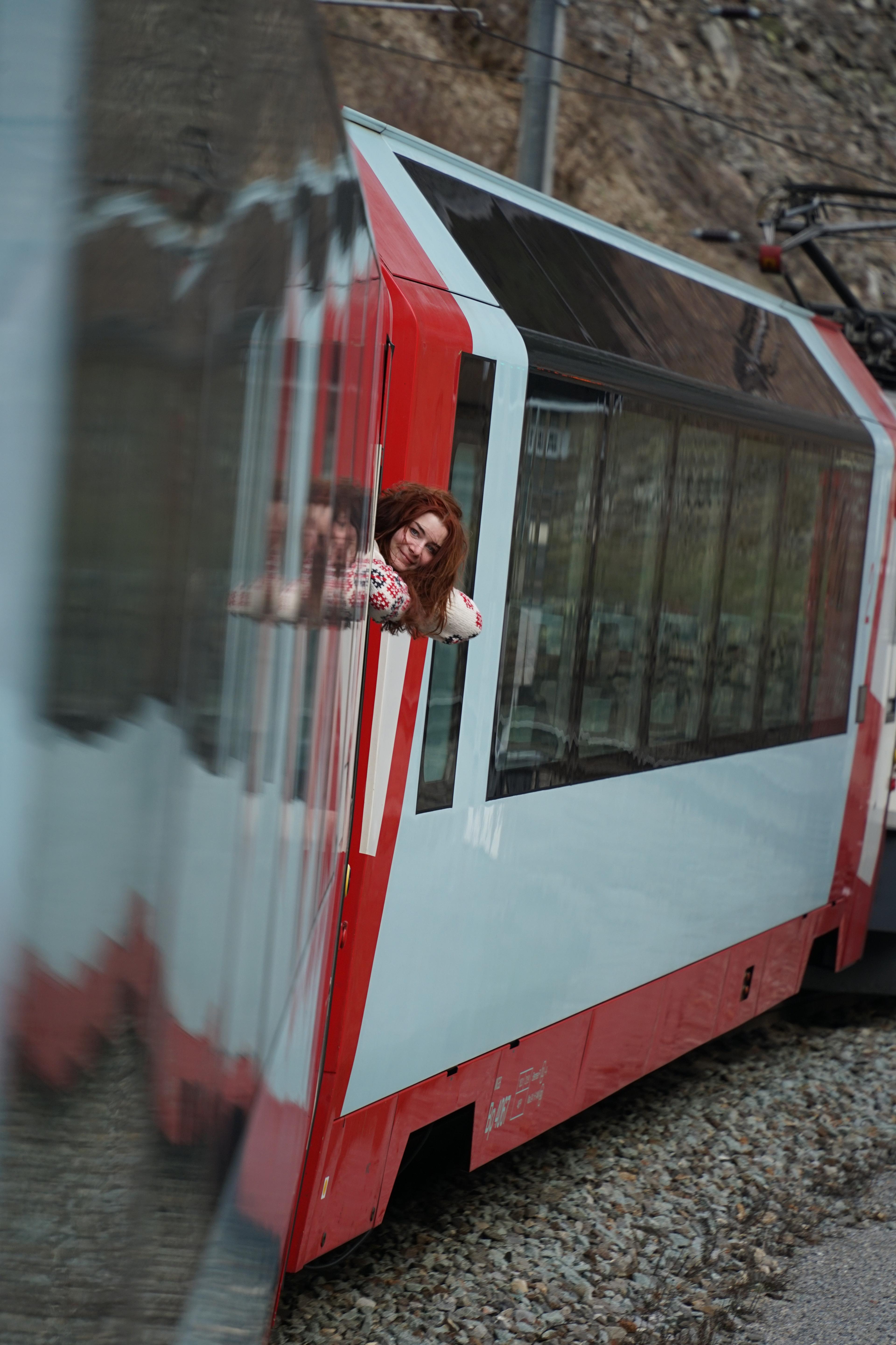 A person leans out of a modern red and white train window, likely traveling through the Swiss Alps.