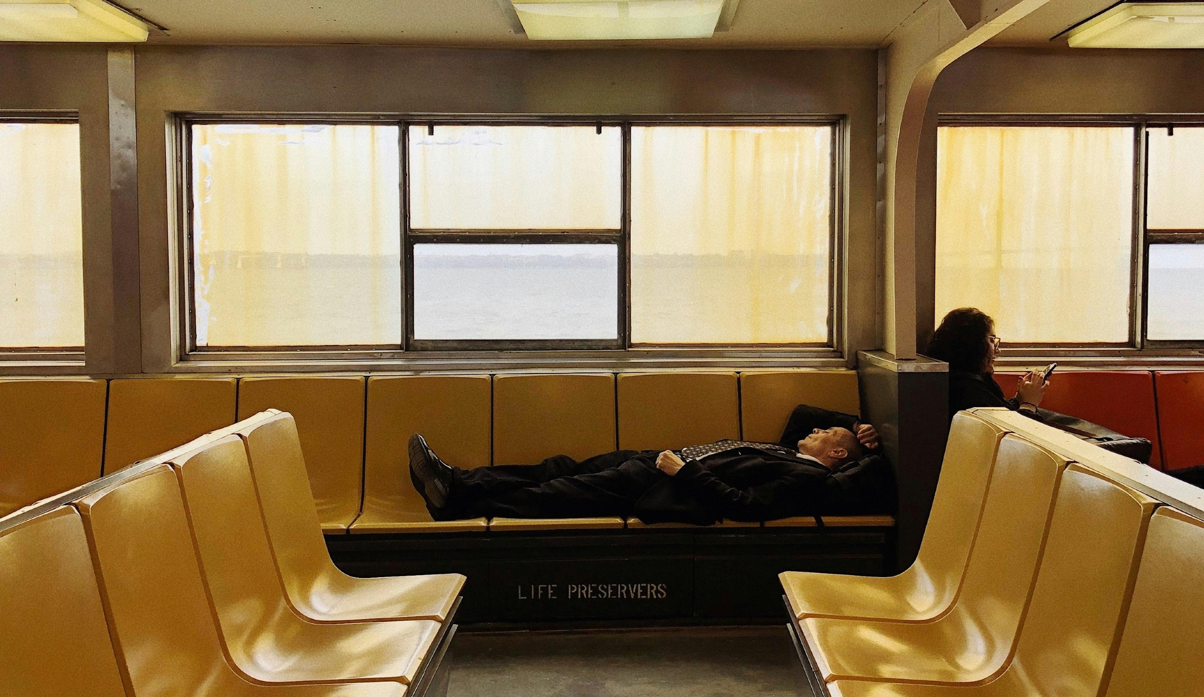 A person rests on yellow seats inside what appears to be a New York City ferry, with "LIFE PRESERVERS" labeled below.