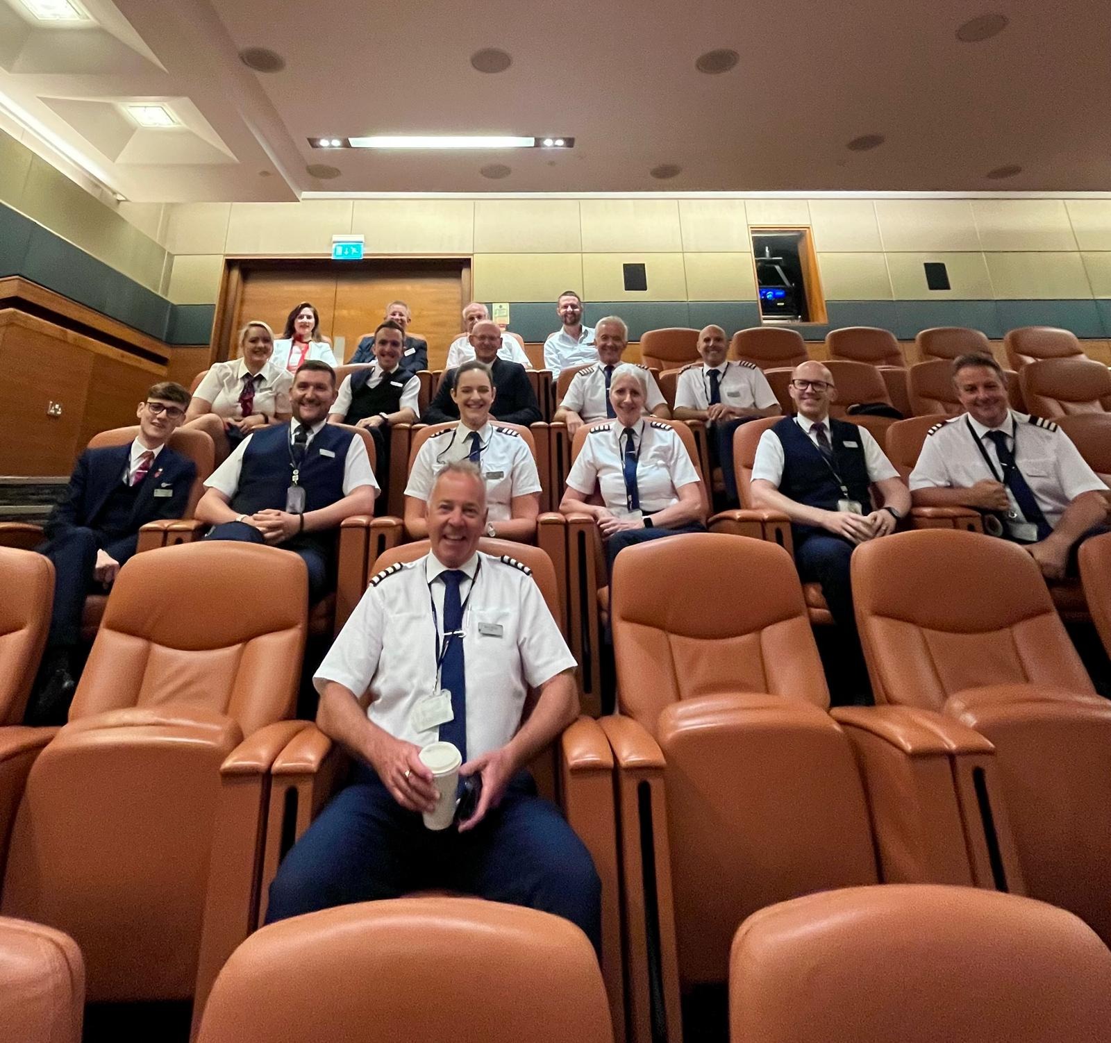 A group of uniformed airline crew members sits together in an auditorium setting, possibly at an airport conference room.