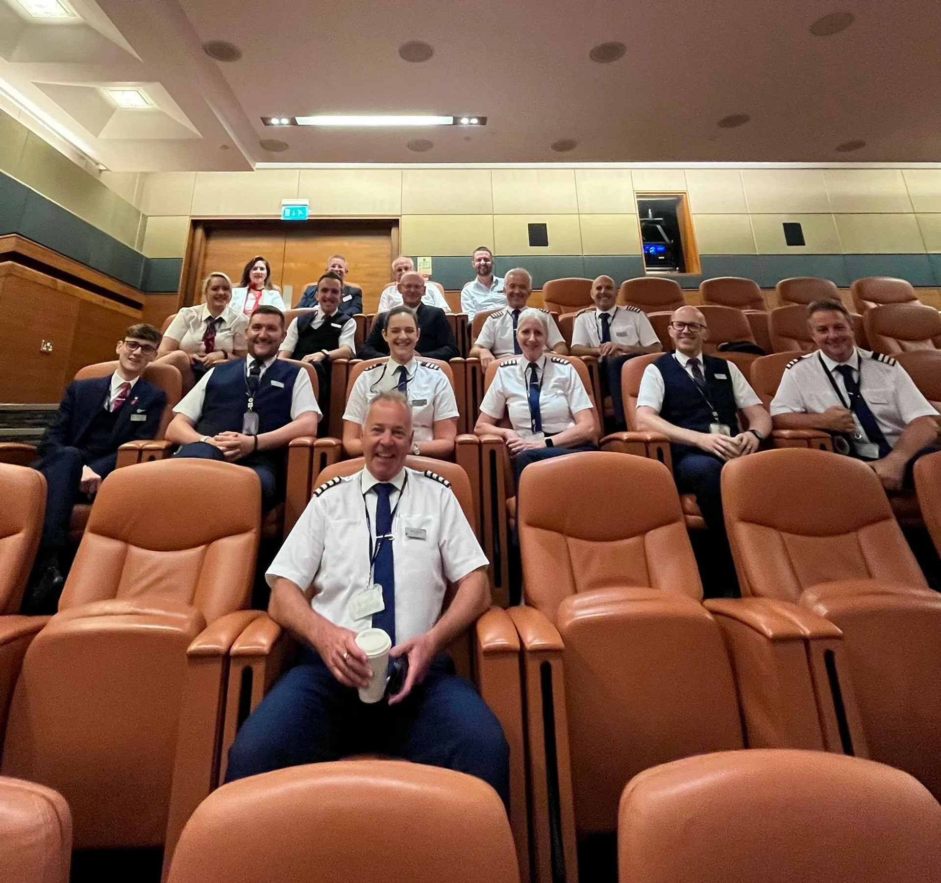 A group of uniformed airline crew members sits together in an auditorium setting, possibly at an airport conference room.