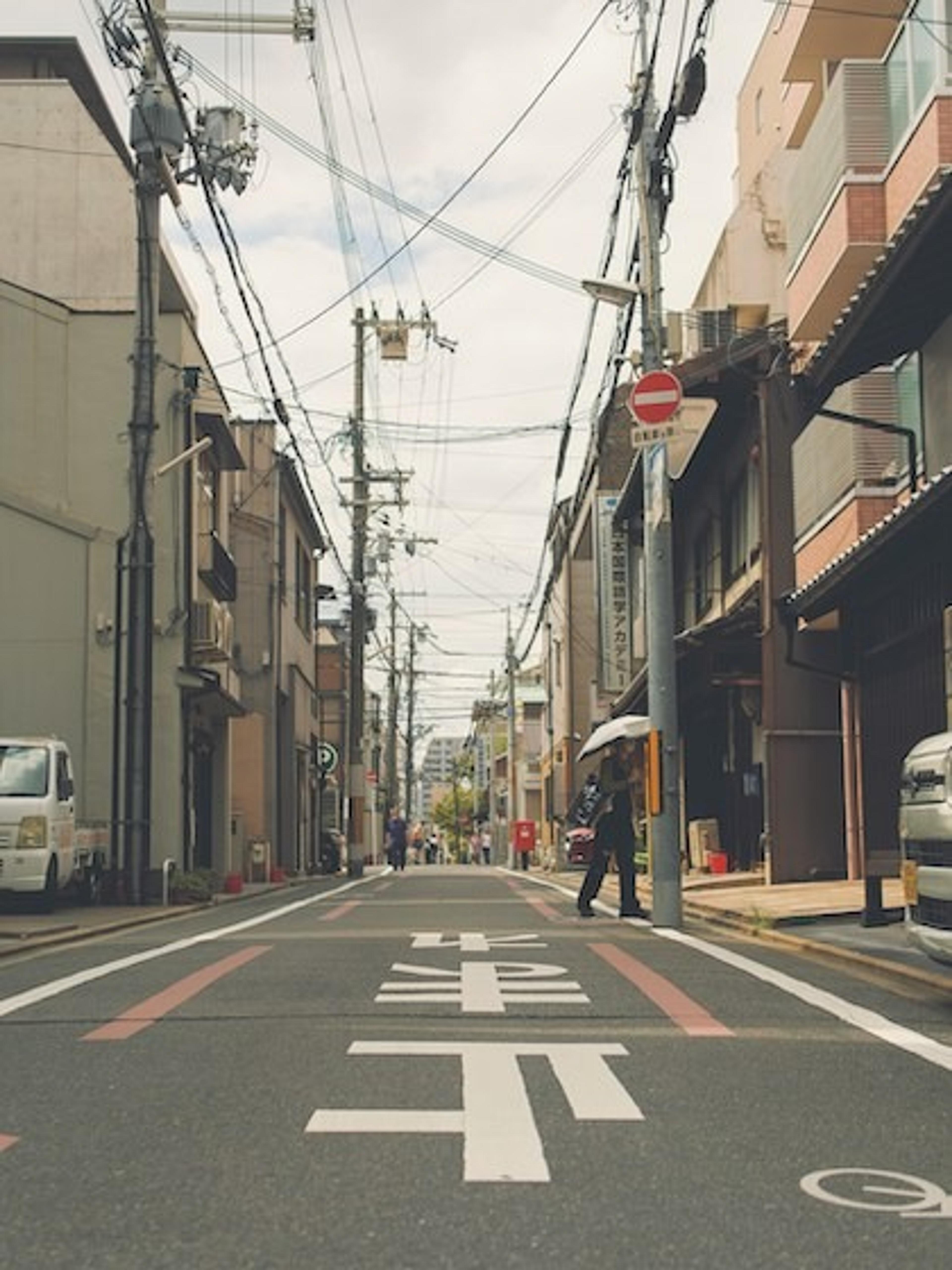 A narrow street in a Japanese city, likely Kyoto, lined with traditional buildings and overhead power lines.