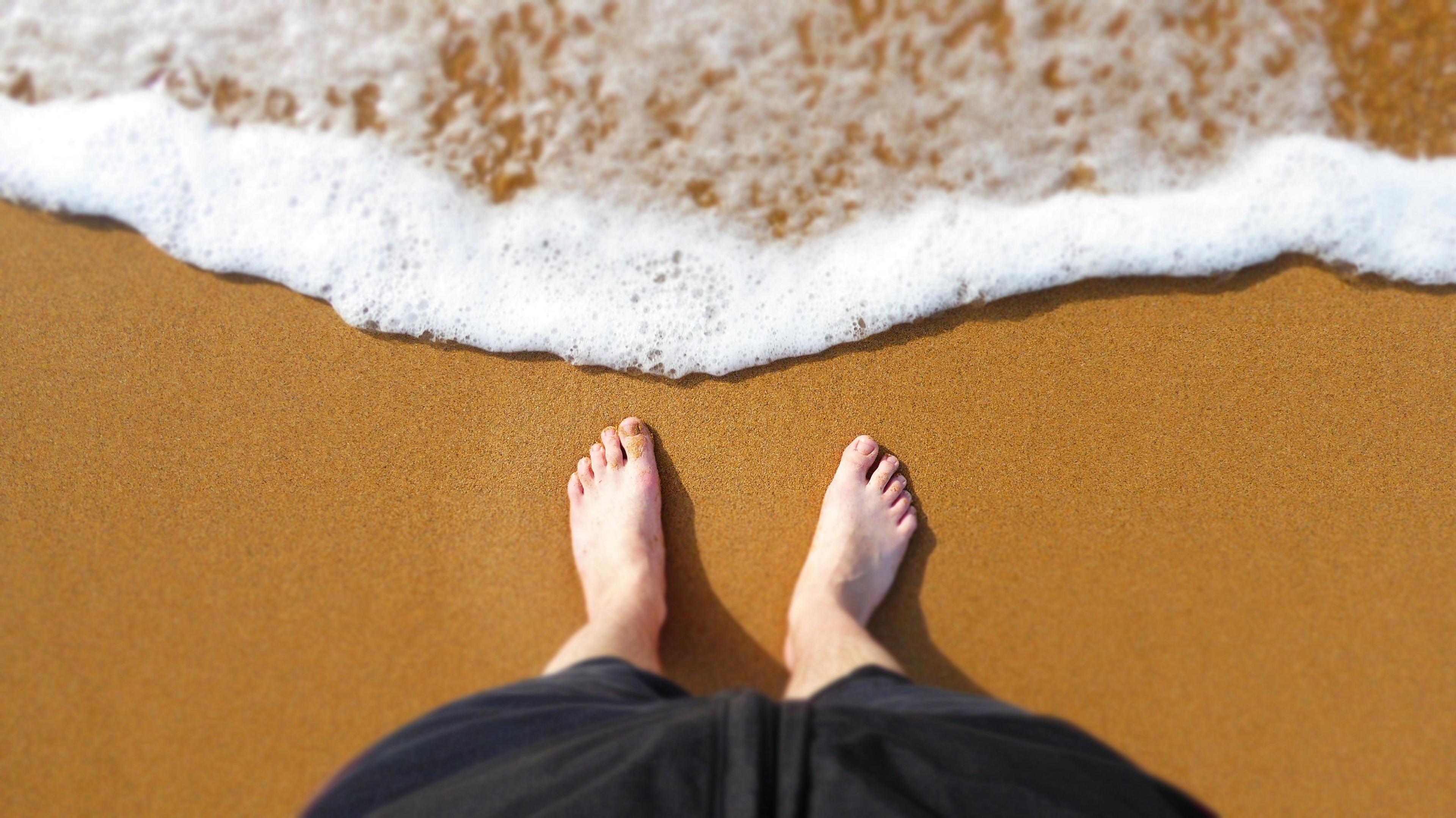 Bare feet stand on the sandy shore as a gentle wave approaches