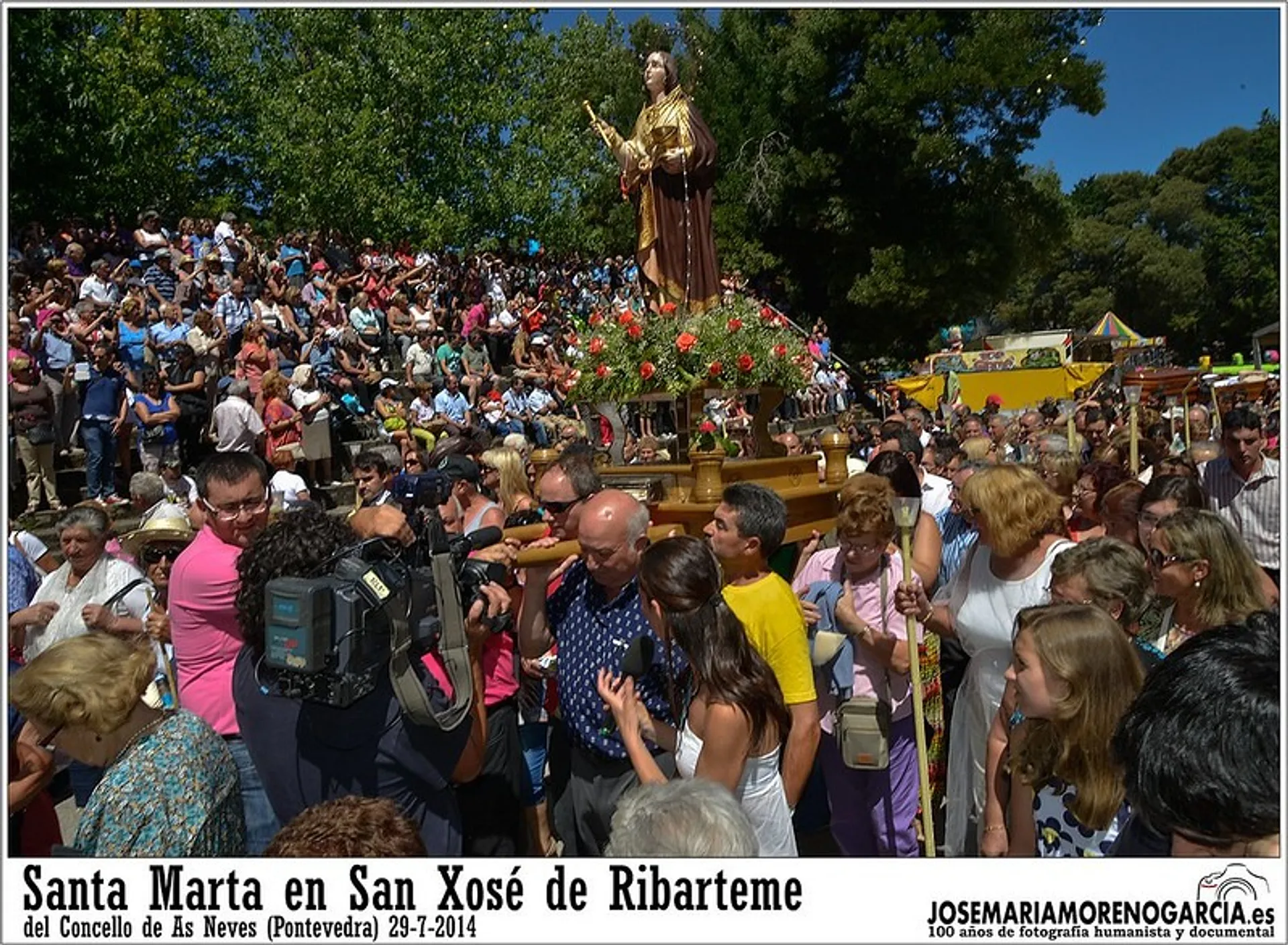 A festive crowd gathers around a statue adorned with flowers in San Xosé de Ribarteme, Pontevedra.
