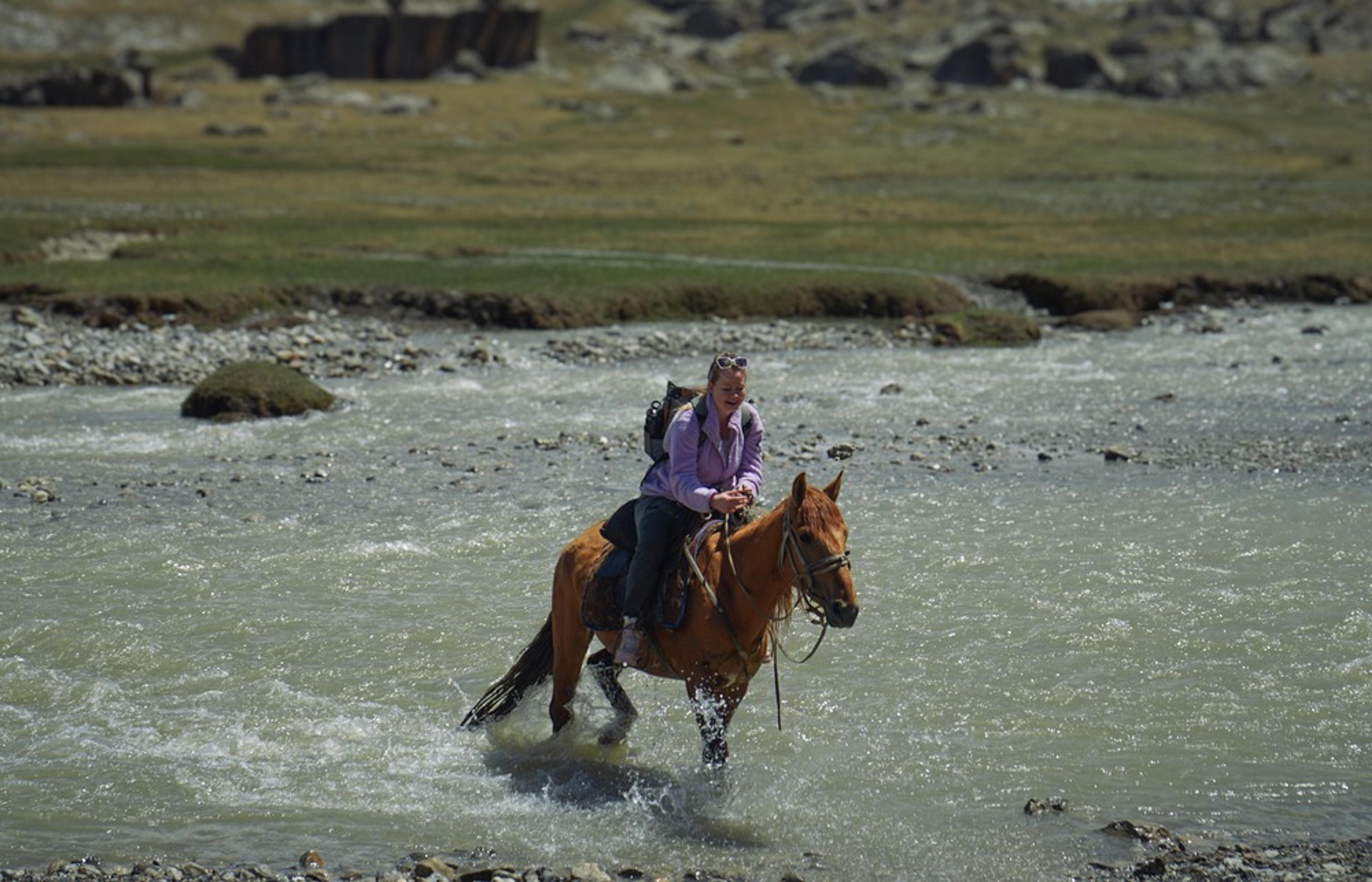 Flight Finder Kristi rides a horse through a shallow river in a mountainous landscape near Kel-Suu Lake, Kyrgyzstan.