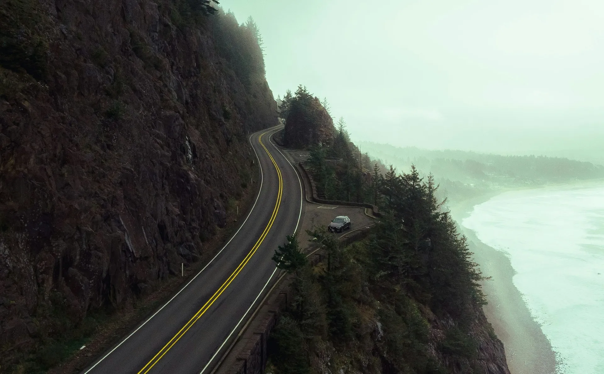 A coastal highway winds along a cliffside in Oregon, overlooking a misty ocean and dense forest.