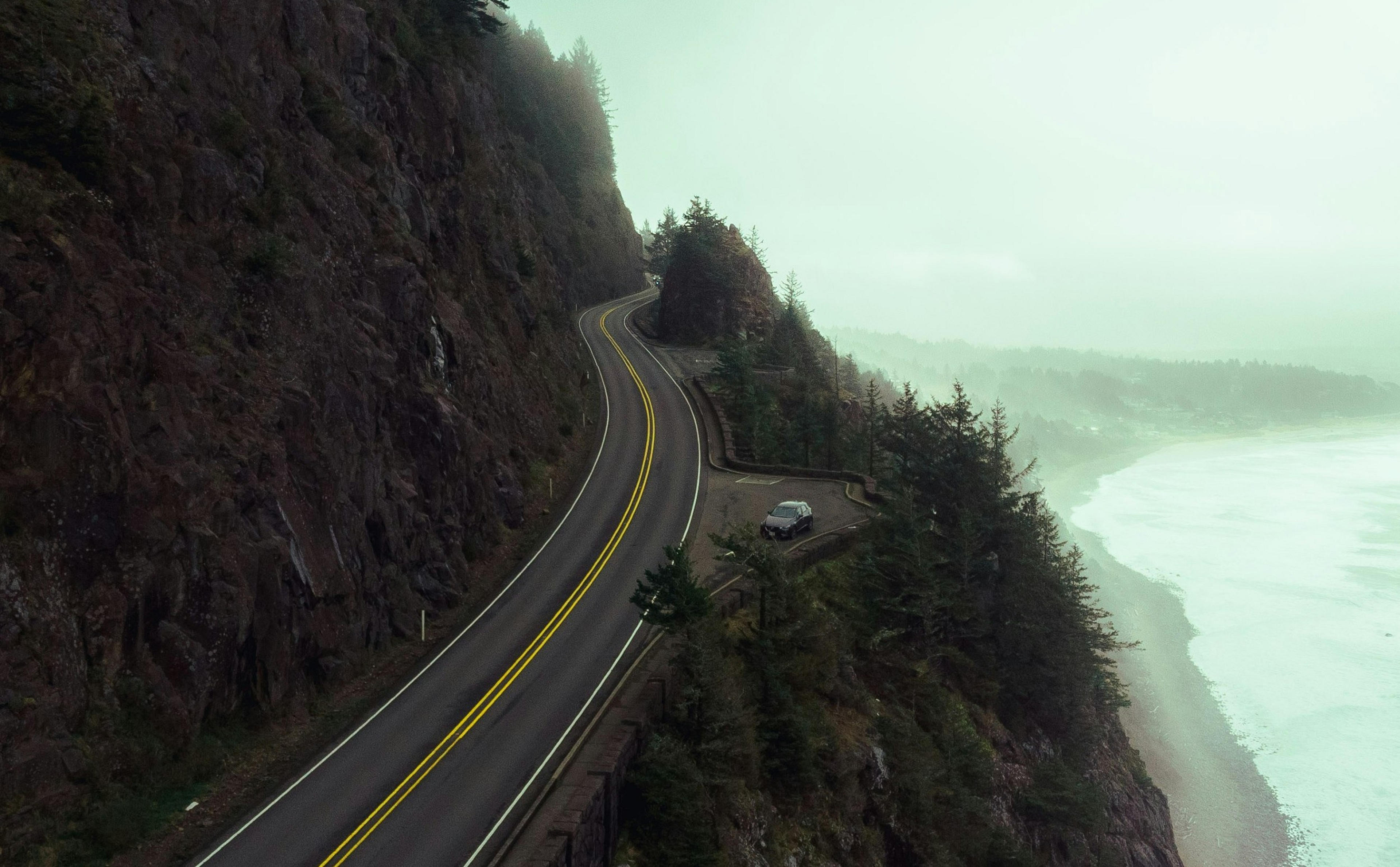 A coastal highway winds along a cliffside in Oregon, overlooking a misty ocean and dense forest.