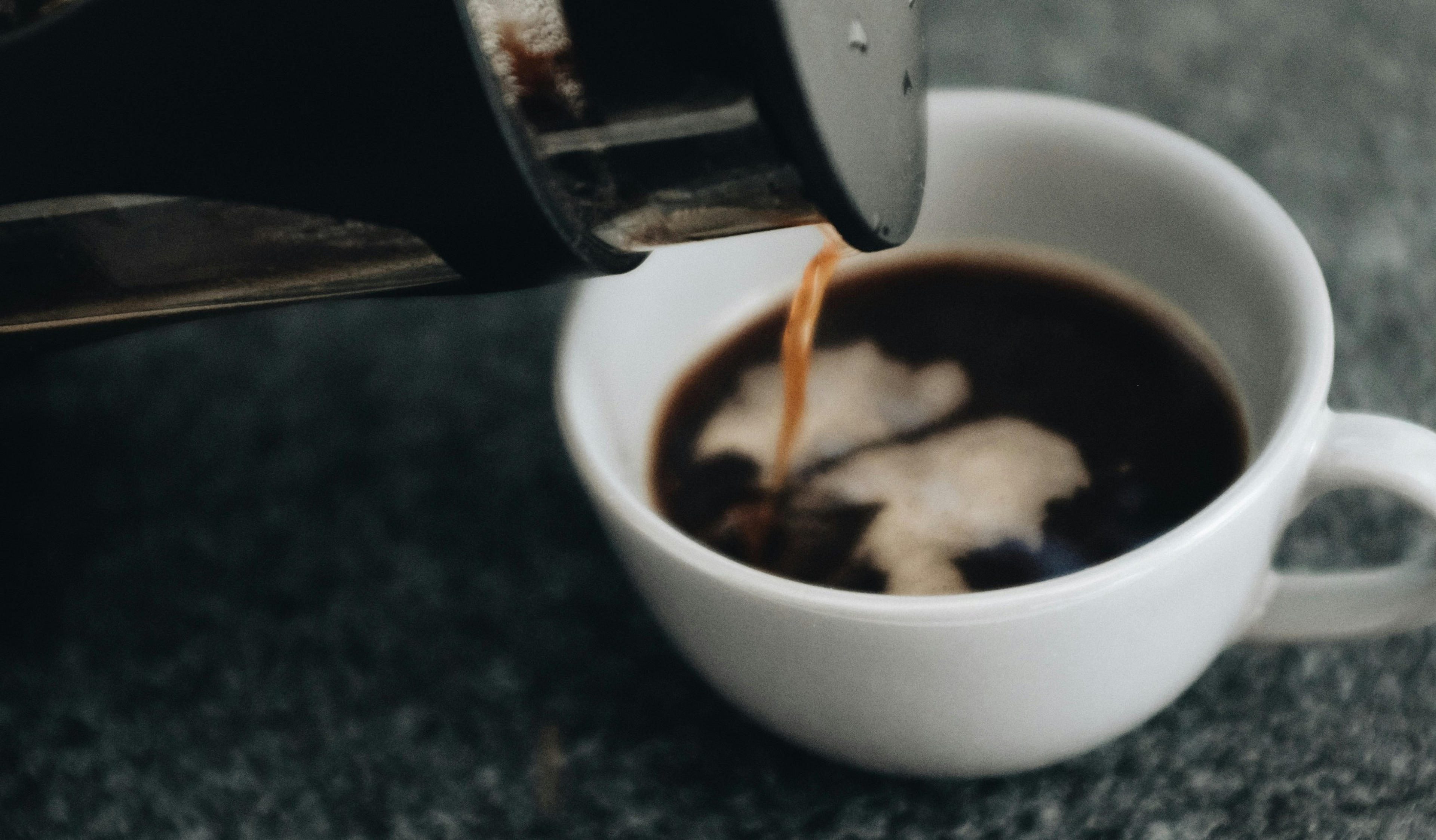A stream of coffee is poured into a white cup on a textured gray surface.