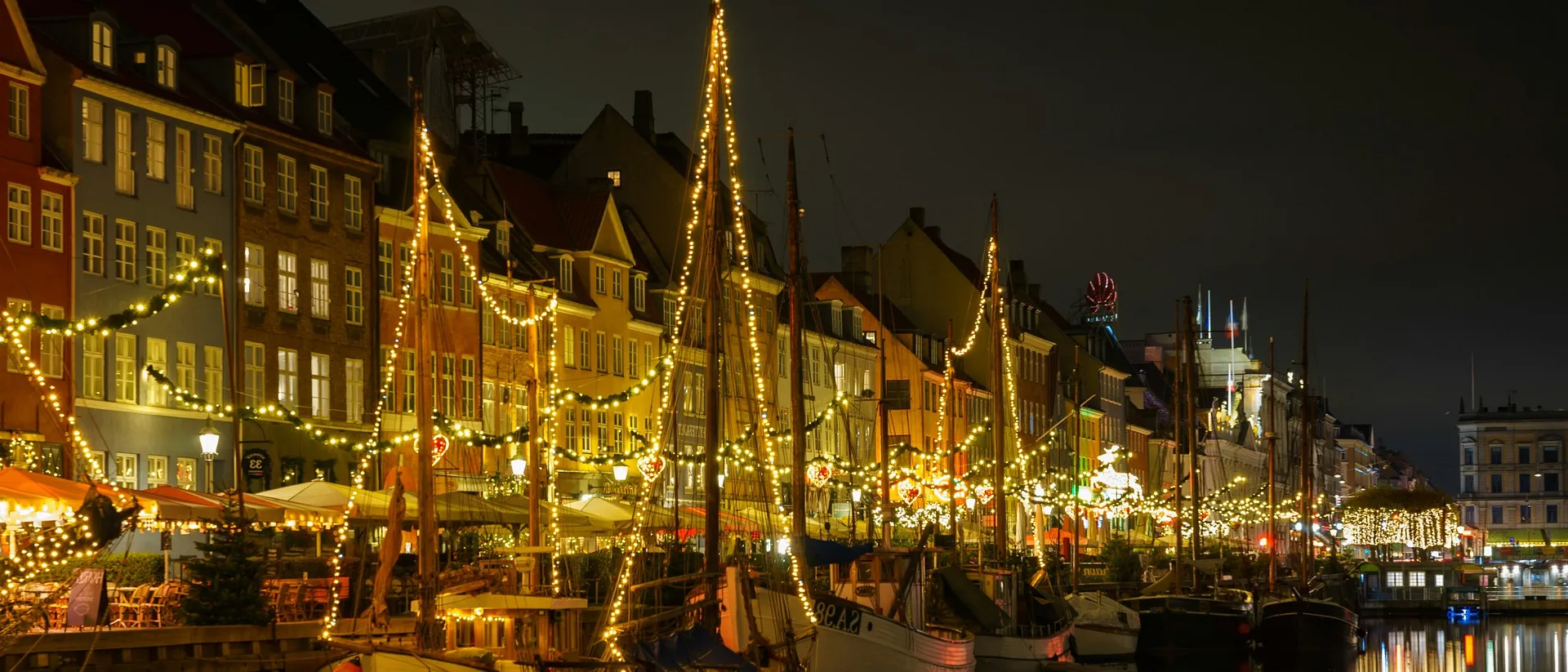 A festive evening scene of the iconic Nyhavn harbor in Copenhagen, Denmark, illuminated with string lights.