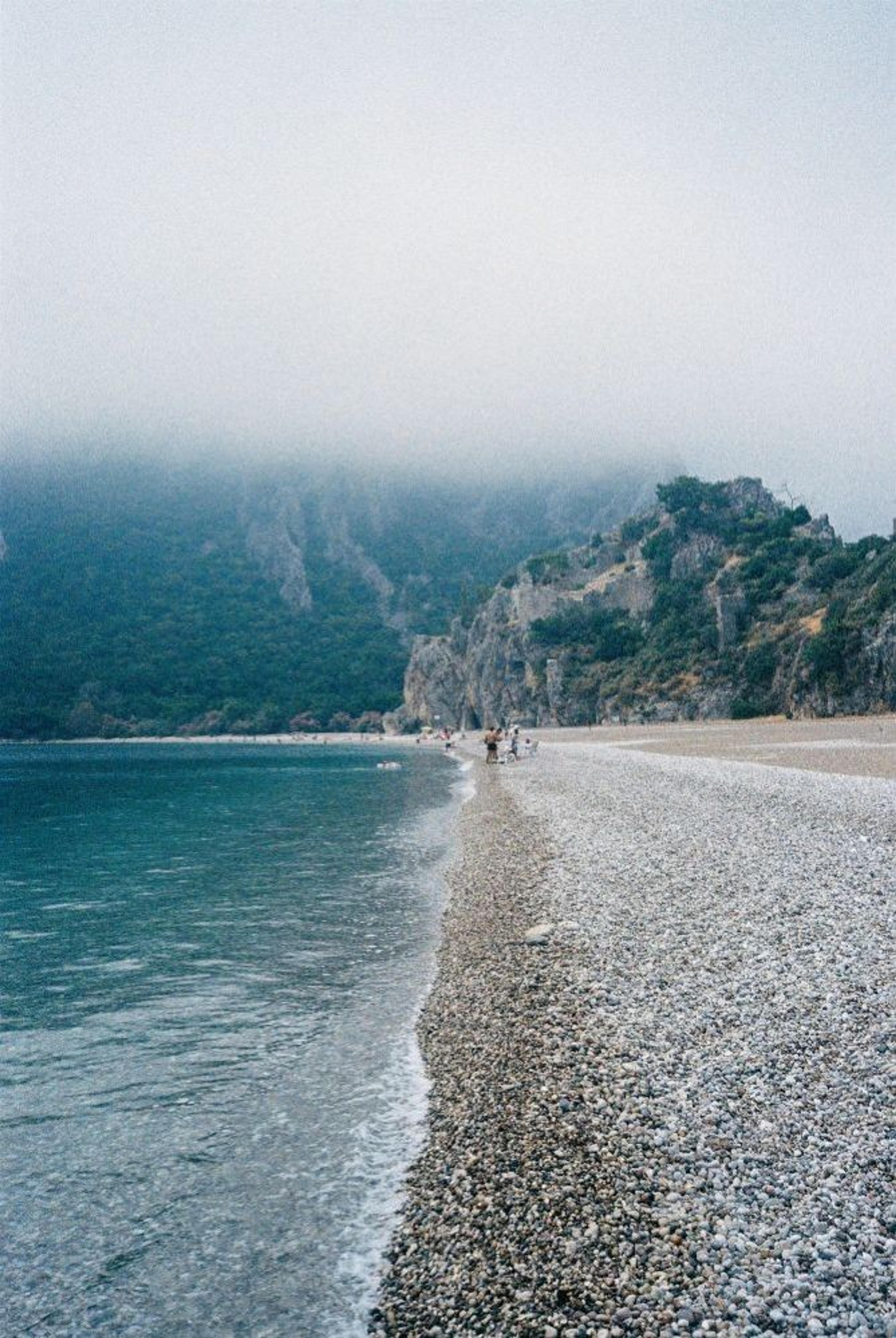 A misty view of Olympos Beach in Turkey, featuring a pebbled shoreline meeting a calm sea under the looming cliffs.