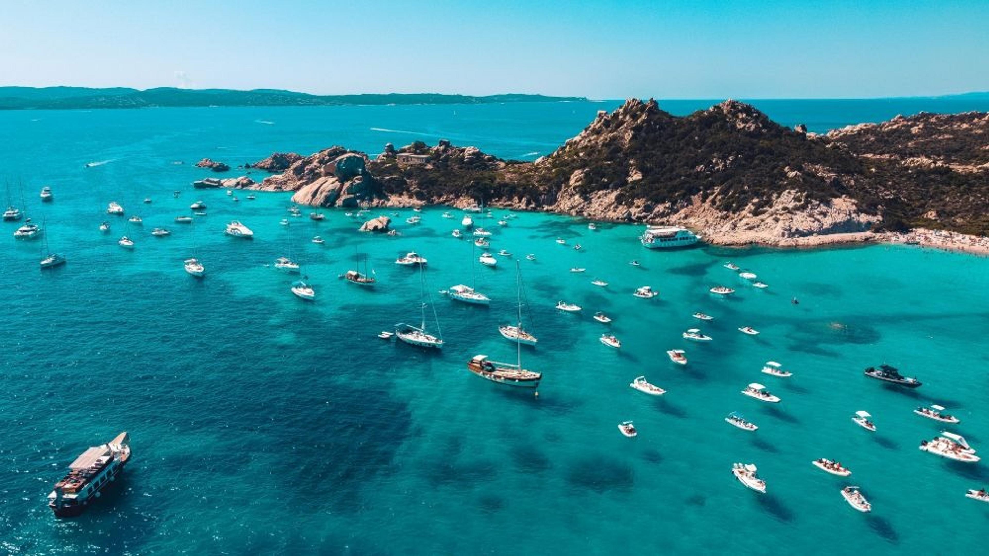 Numerous boats float on the turquoise waters surrounding the rocky shores of La Maddalena Archipelago in Sardinia.