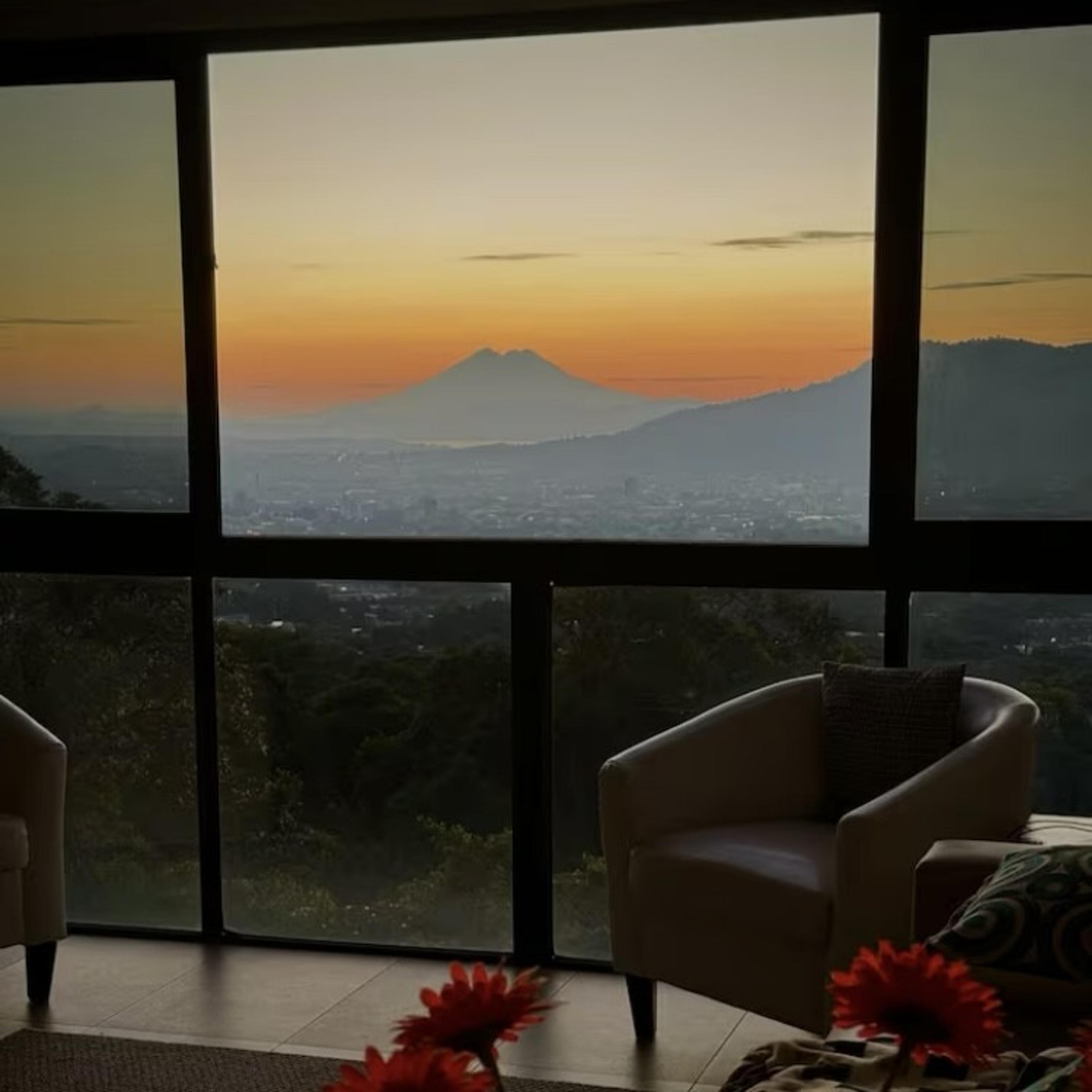 A breathtaking view of the Acatenango volcano is visible through a window at sunset from a cozy living space.