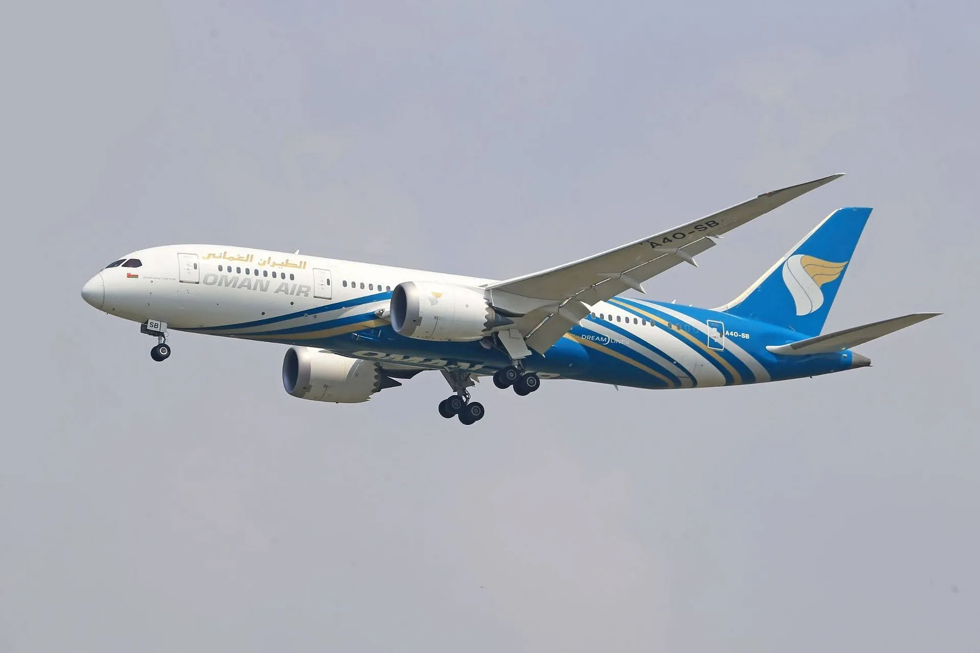 A Boeing 787 Dreamliner from Oman Air, with landing gear extended, glides through a cloudy sky.