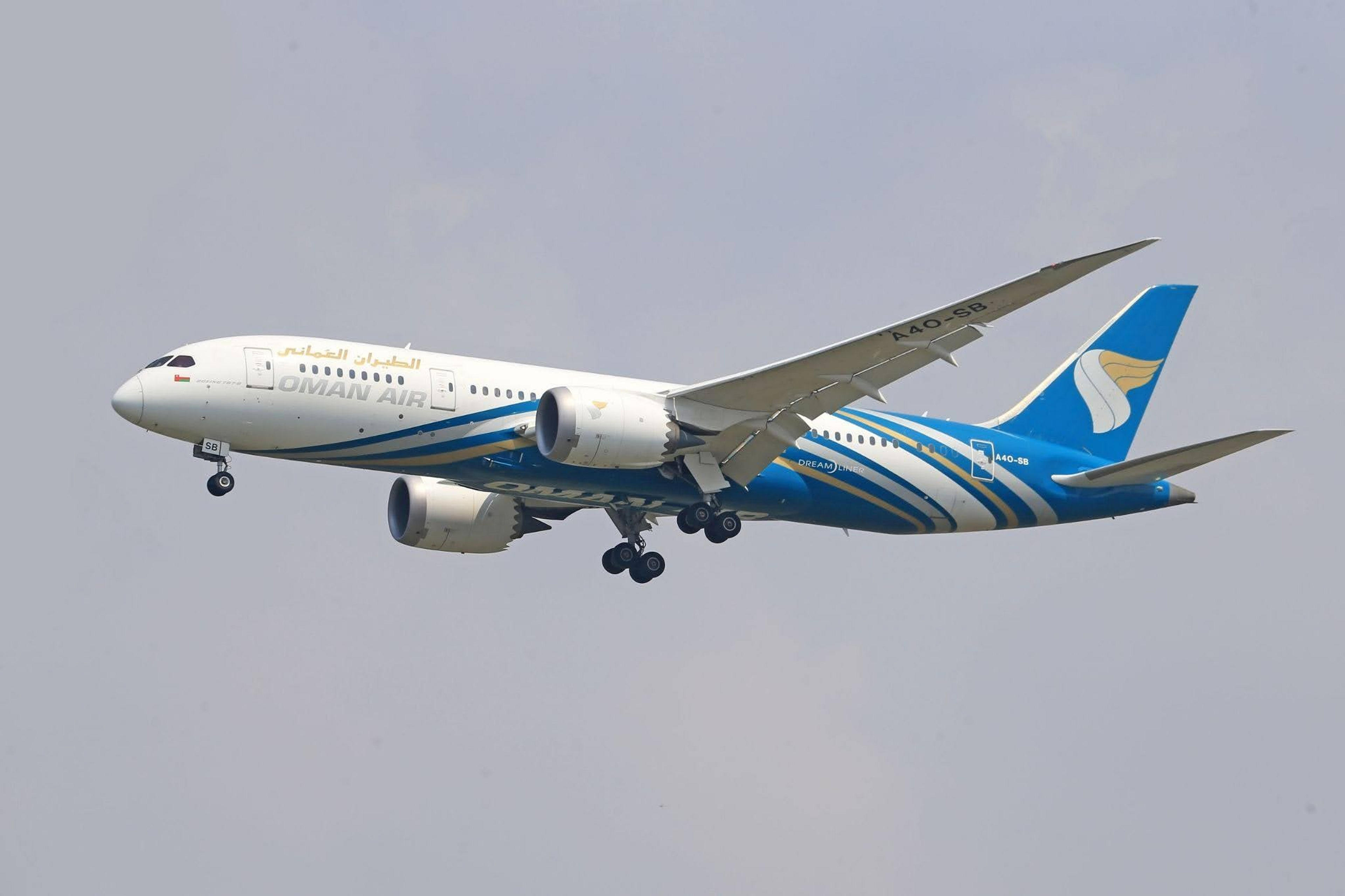 A Boeing 787 Dreamliner from Oman Air, with landing gear extended, glides through a cloudy sky.