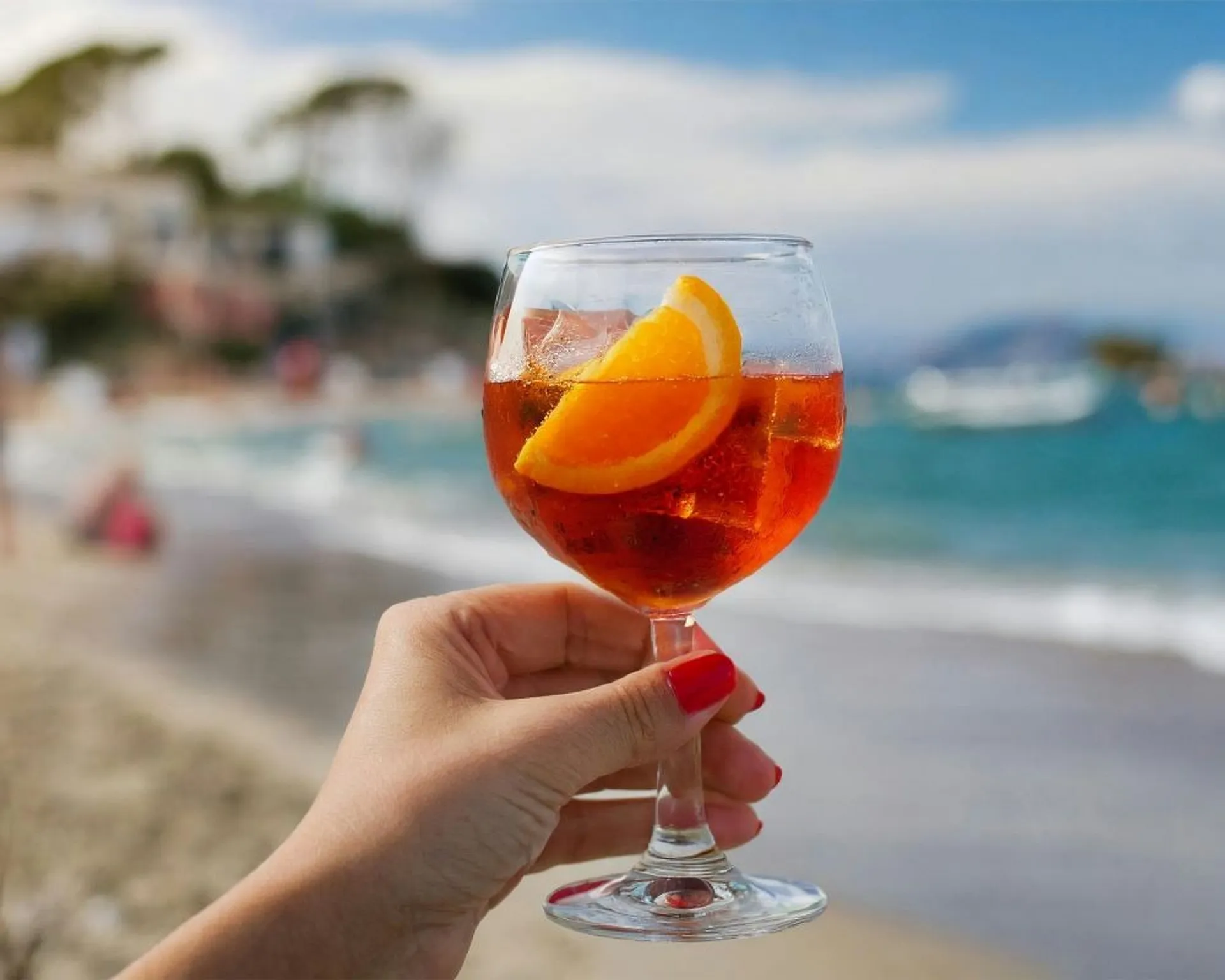 A hand holding a glass of Aperol Spritz on a beach, likely in a Mediterranean setting.