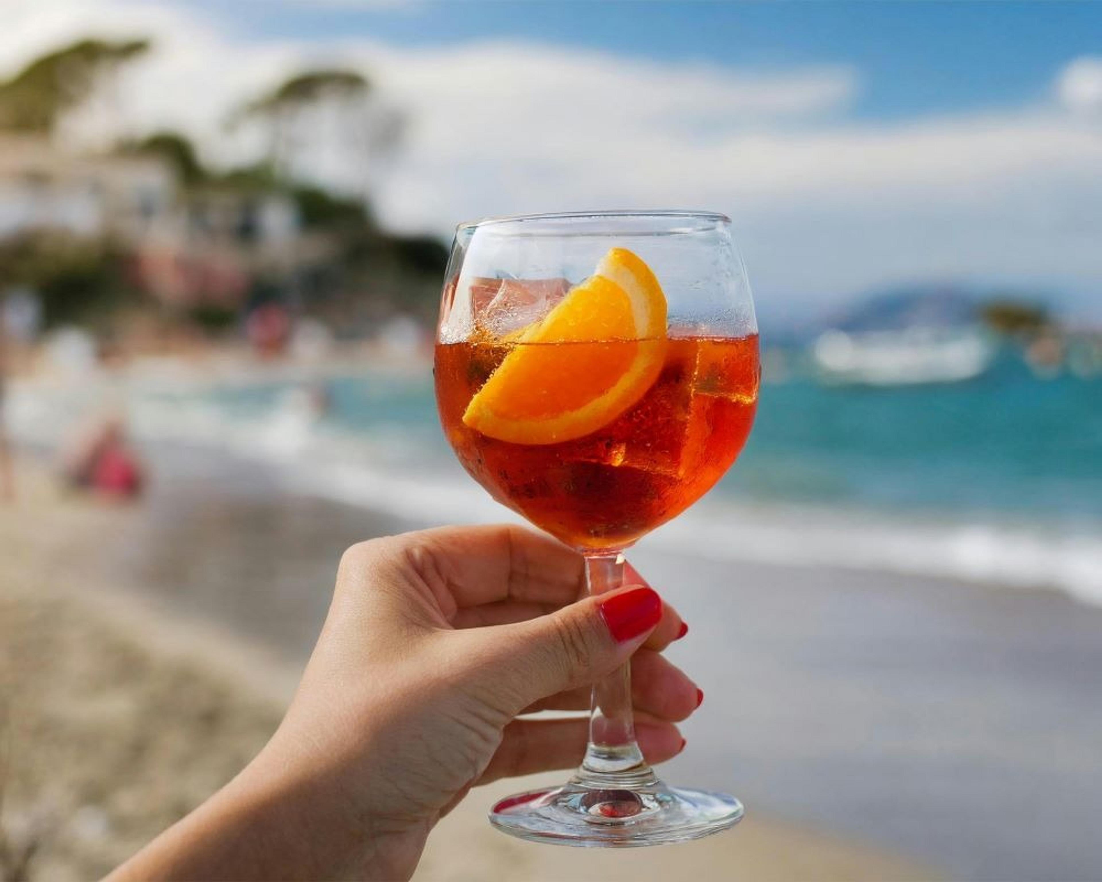 A hand holding a glass of Aperol Spritz on a beach, likely in a Mediterranean setting.