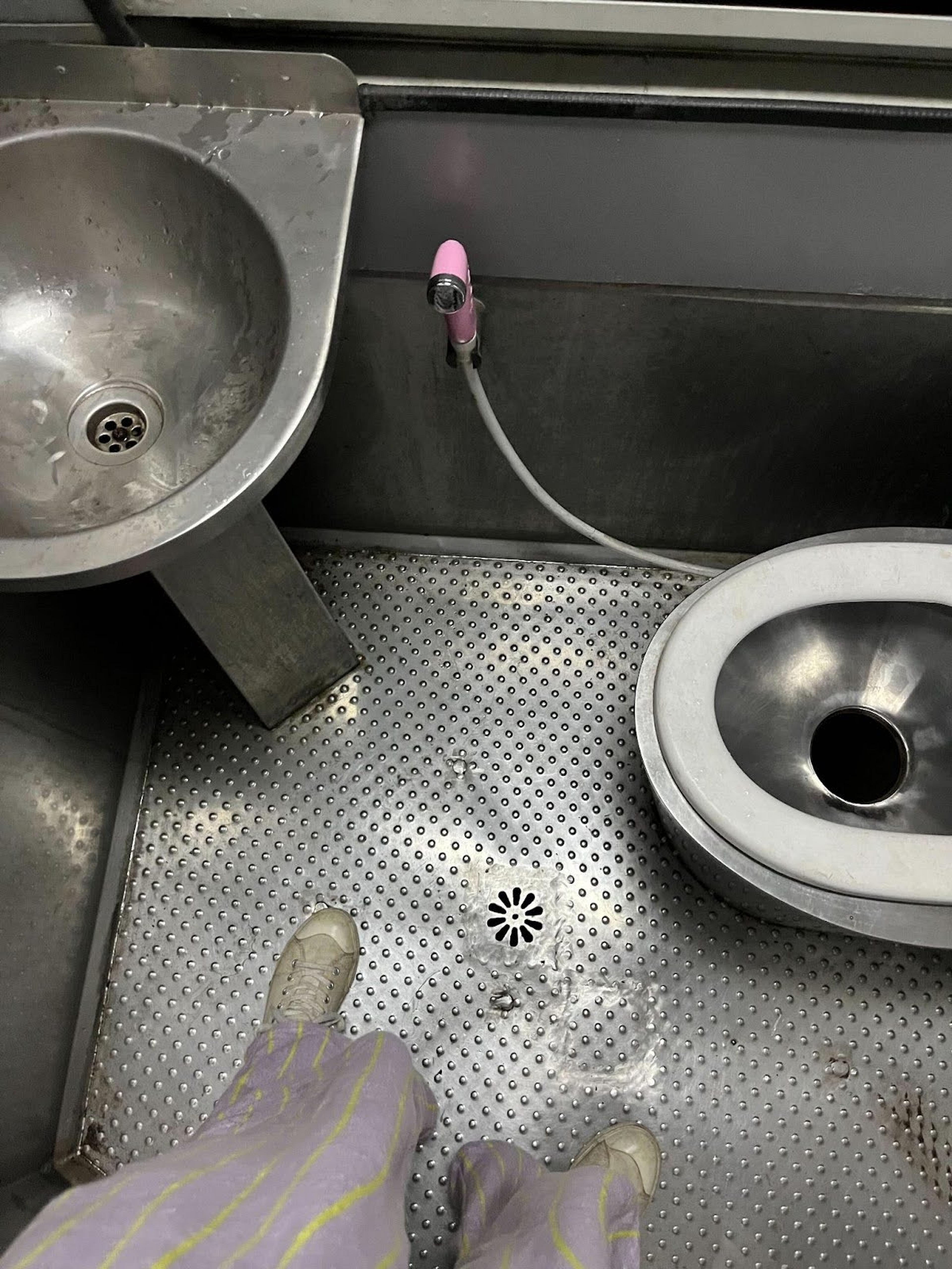 A stainless steel bathroom in a train compartment with a toilet, sink, and hose.