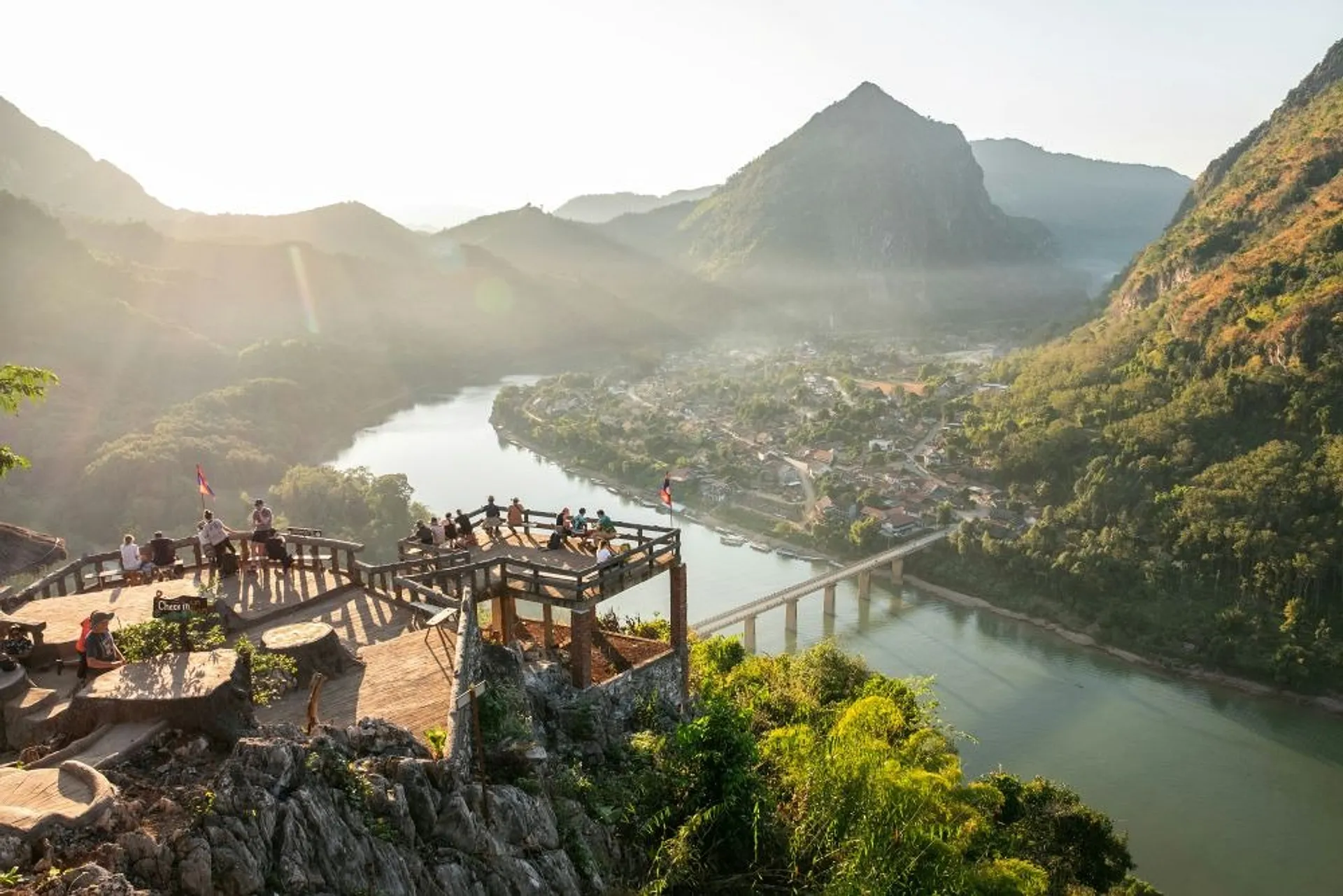 A scenic viewpoint overlooking the Nam Song River and lush mountains in Vang Vieng, Laos, captures a peaceful afternoon with visitors enjoying the view.