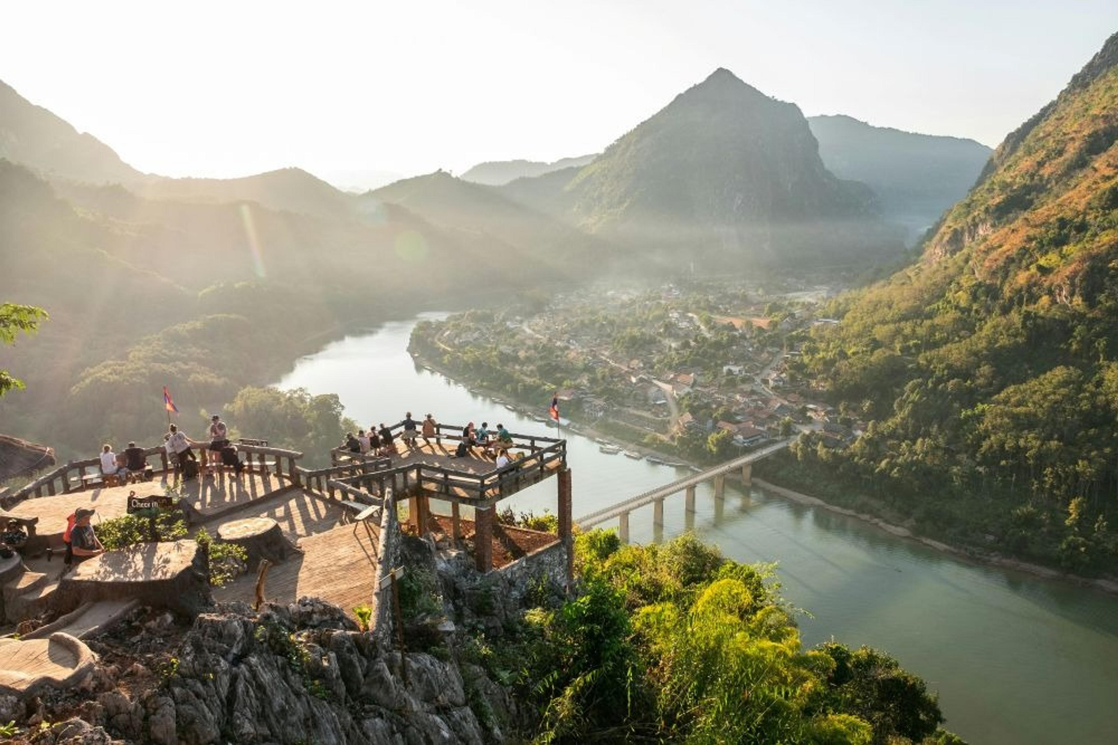 A scenic viewpoint overlooking the Nam Song River and lush mountains in Vang Vieng, Laos, captures a peaceful afternoon with visitors enjoying the view.