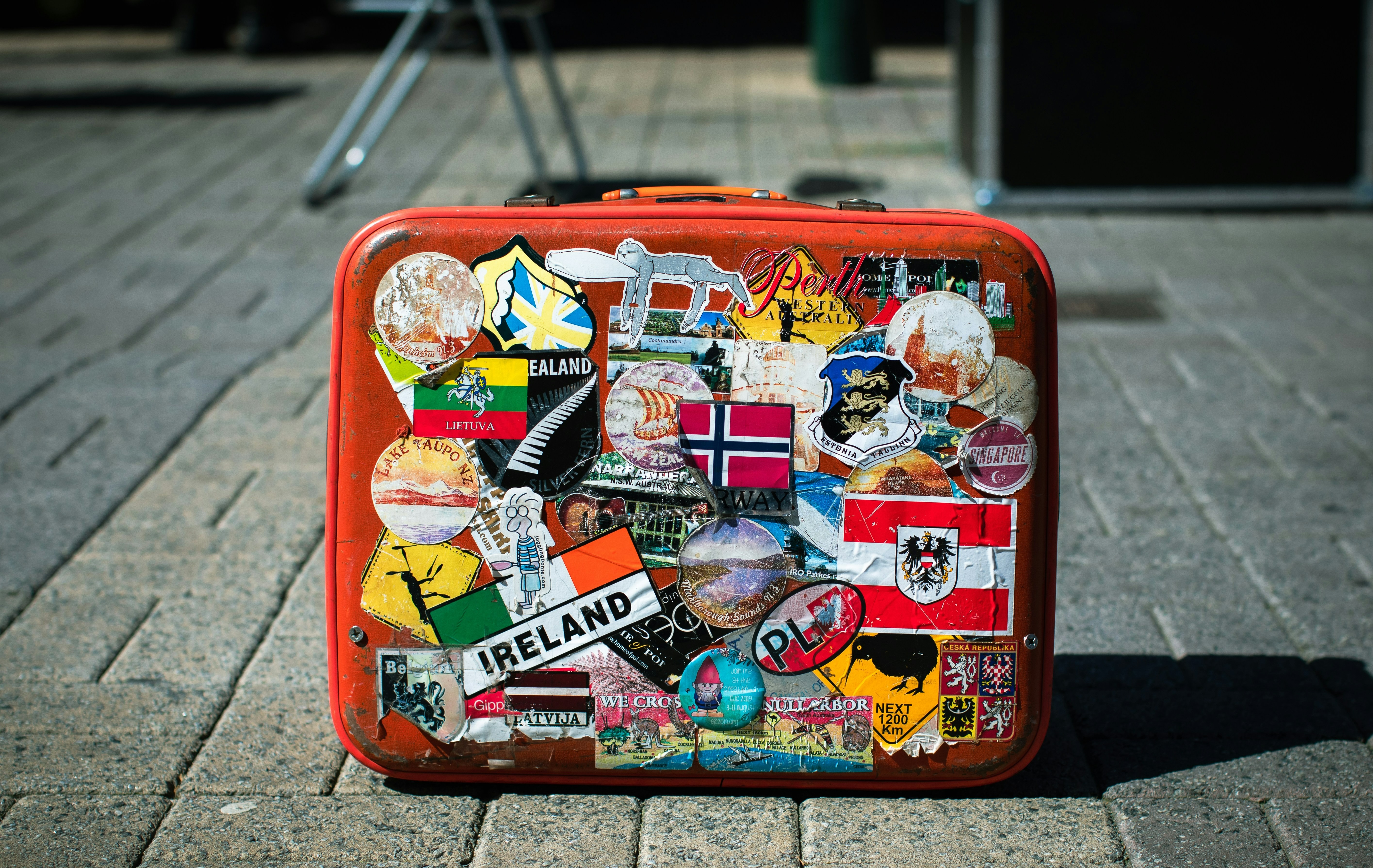A well-traveled suitcase, adorned with stickers from various countries including Ireland and Norway, stands on a stone pavement.