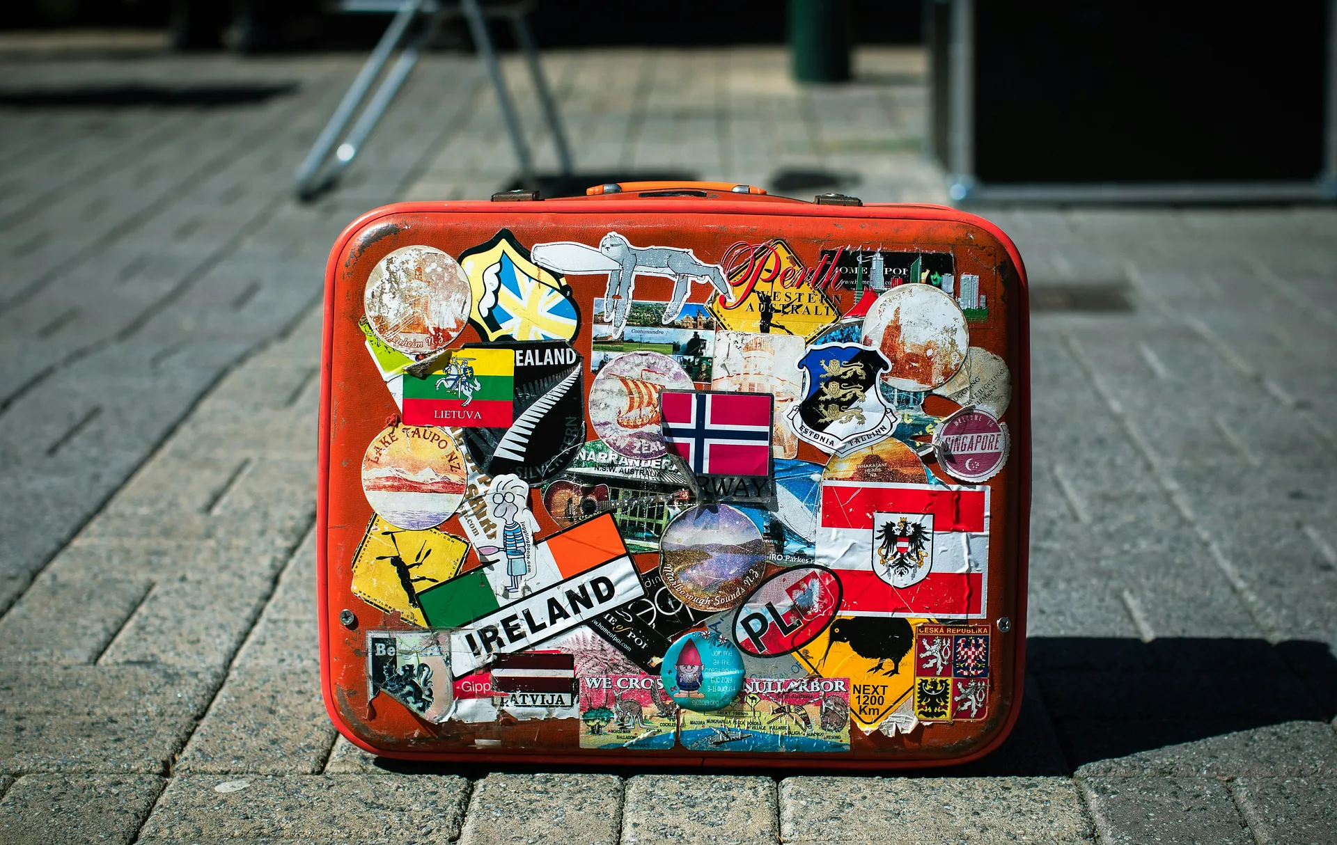 A well-traveled suitcase, adorned with stickers from various countries including Ireland and Norway, stands on a stone pavement.