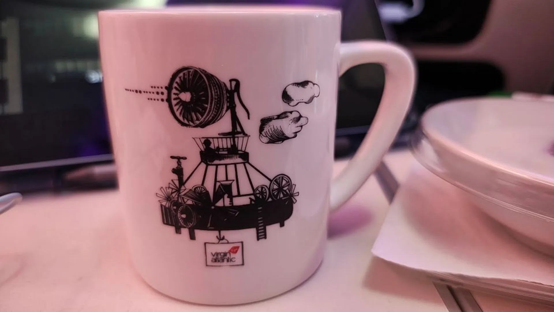 A Virgin Atlantic mug featuring artistic sketches of mechanical components rests on a surface, likely inside an aircraft cabin.