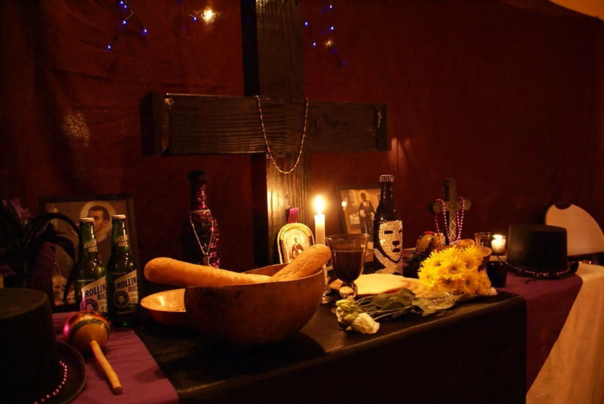A dimly lit altar is adorned with various offerings including candles, flowers, a wooden cross, and bottles in a setting that appears to be part of a religious or cultural ritual, possibly in New Orleans.