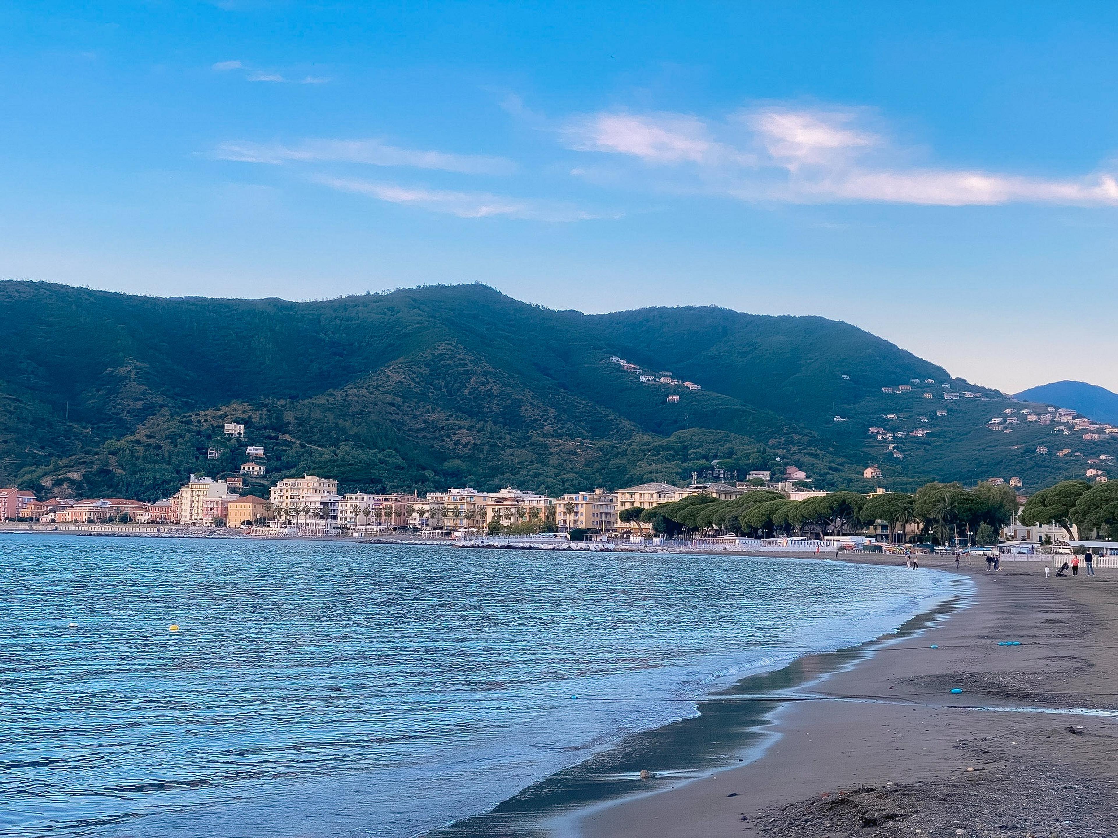 A peaceful beach scene unfolds along the shoreline with lush green hills and charming seaside buildings.