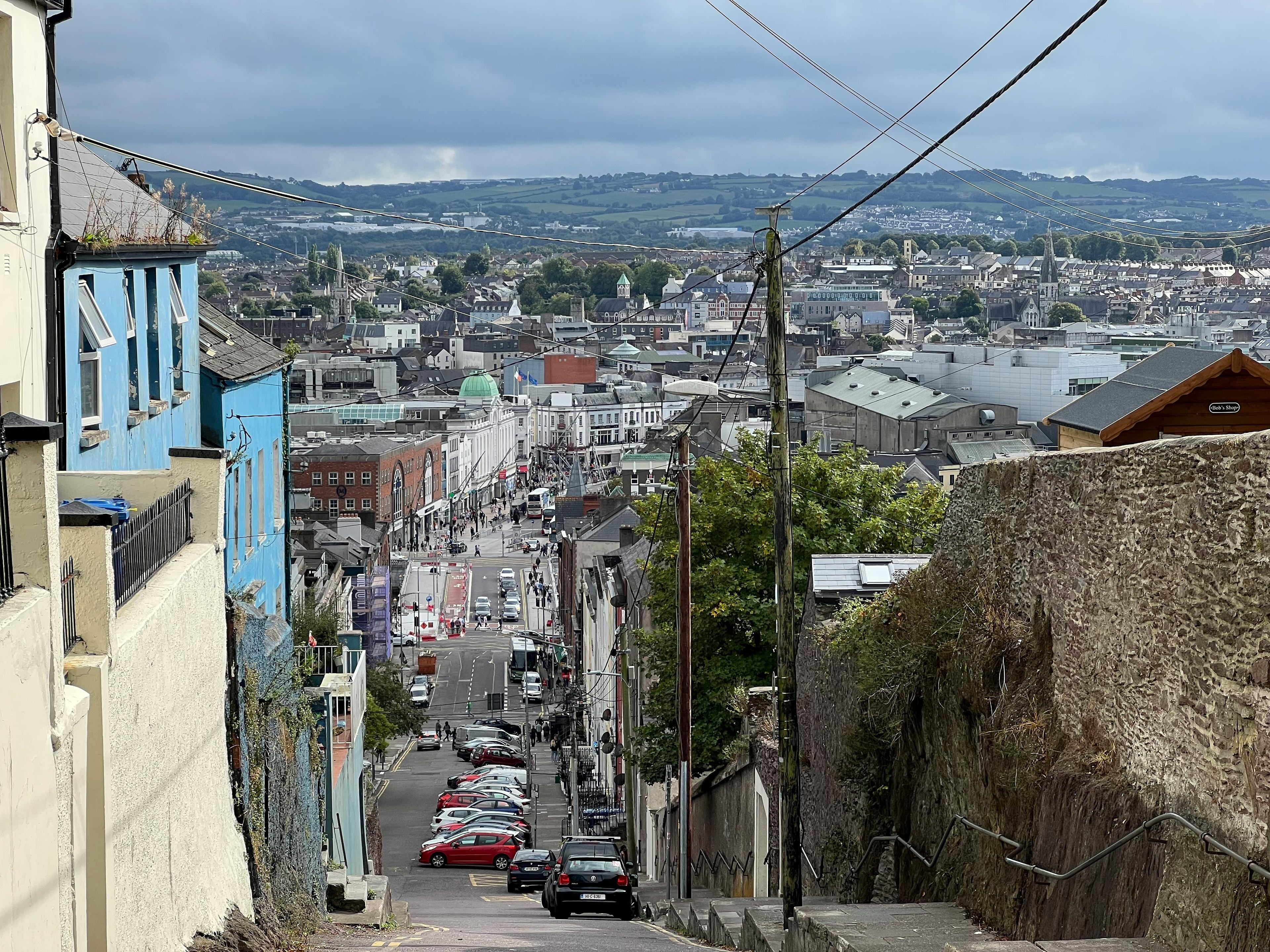 A steep, narrow road descends into the bustling urban landscape of Cork, Ireland, with distant hills visible under a cloudy sky.