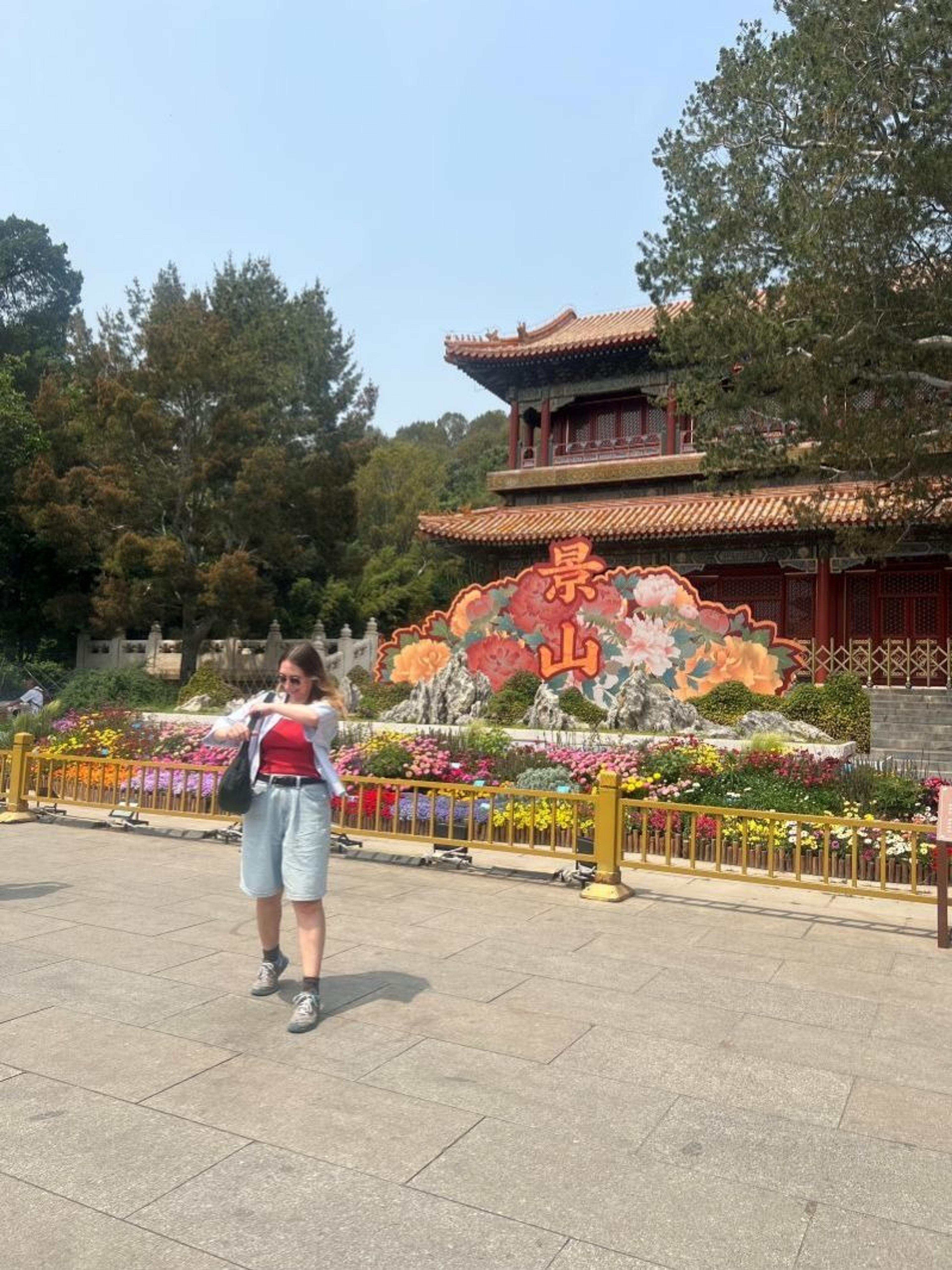 A person stands in front of the intricate entrance to Jing Shan Park, Beijing, surrounded by colorful flowers and traditional Chinese architecture.
