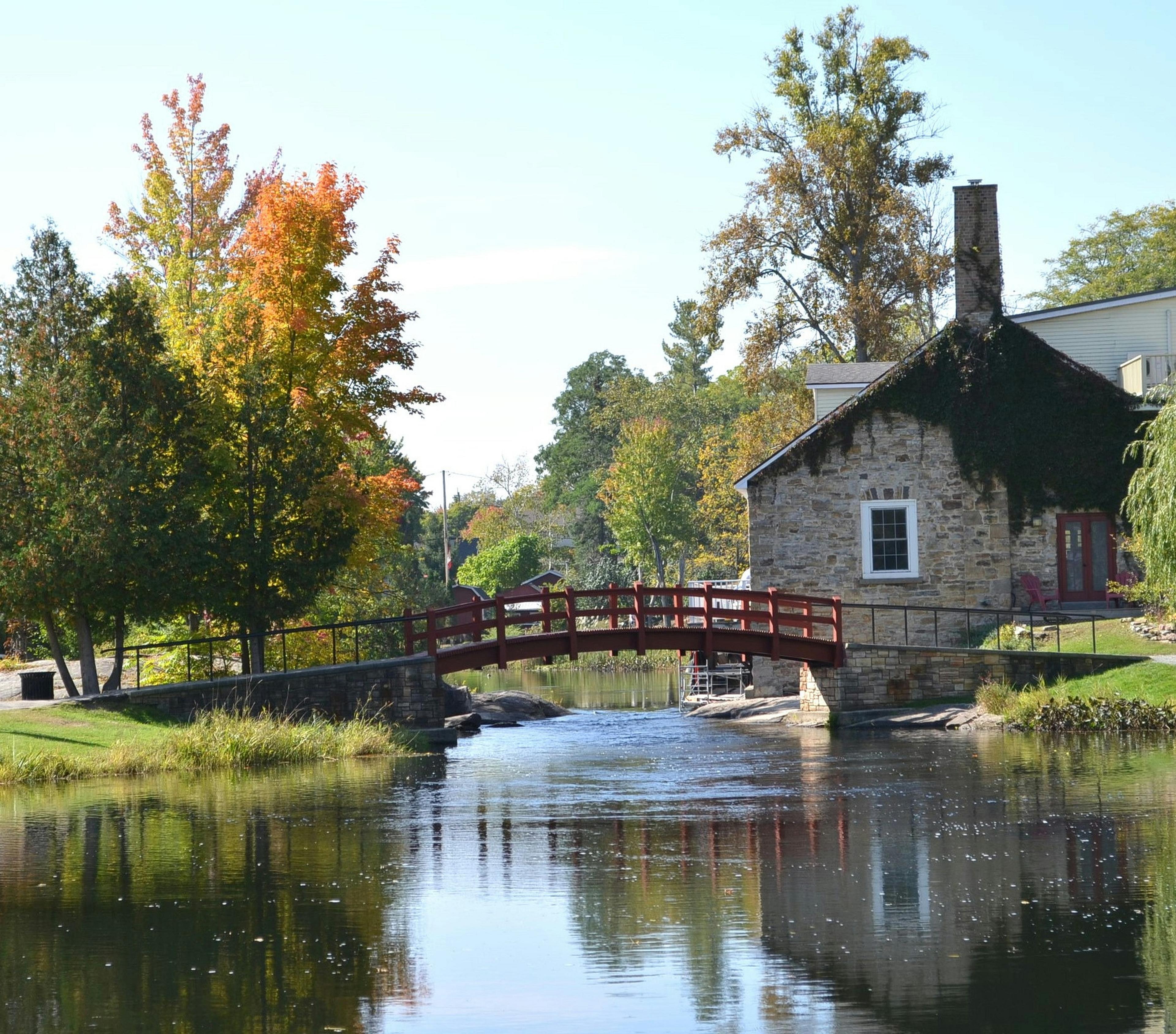 A charming red wooden bridge crosses a tranquil pond near a historic stone building in the picturesque village of Merrickville, Ontario.