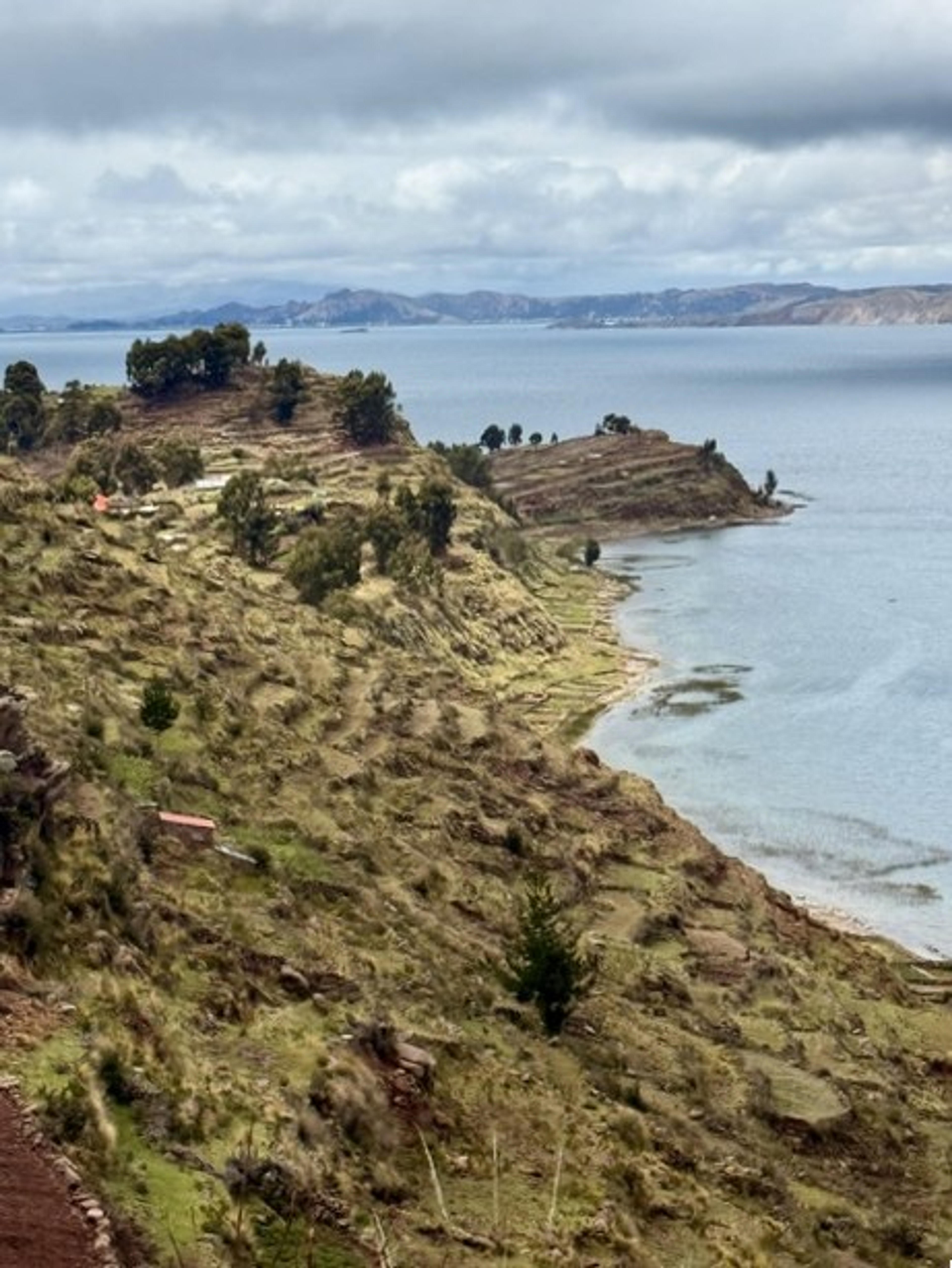 Terraced hills gently slope towards the waters of Lake Titicaca, bordered by a cloudy sky.