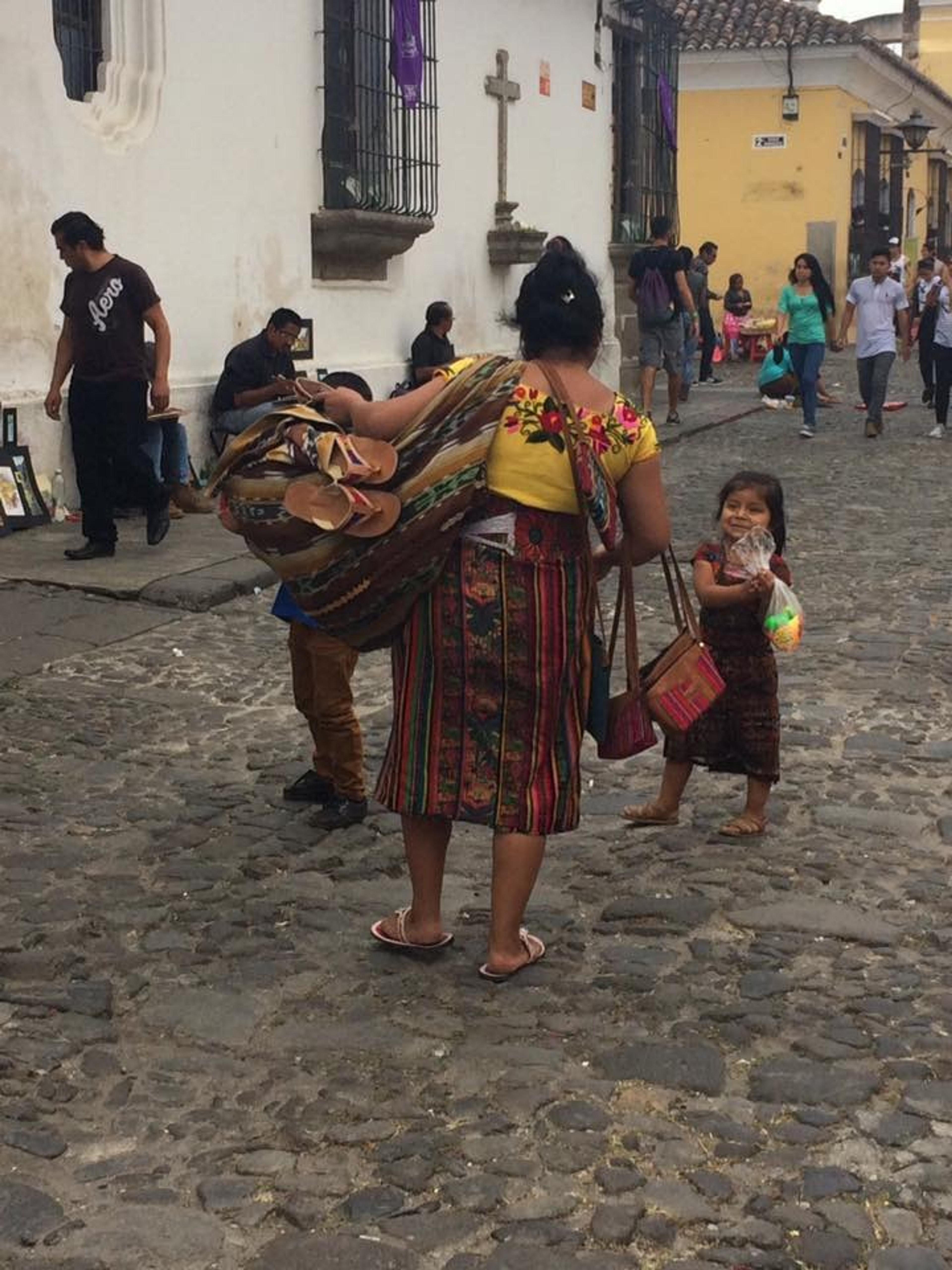 A cobblestone street scene in Antigua, Guatemala features people in traditional attire, including a woman carrying handmade goods.