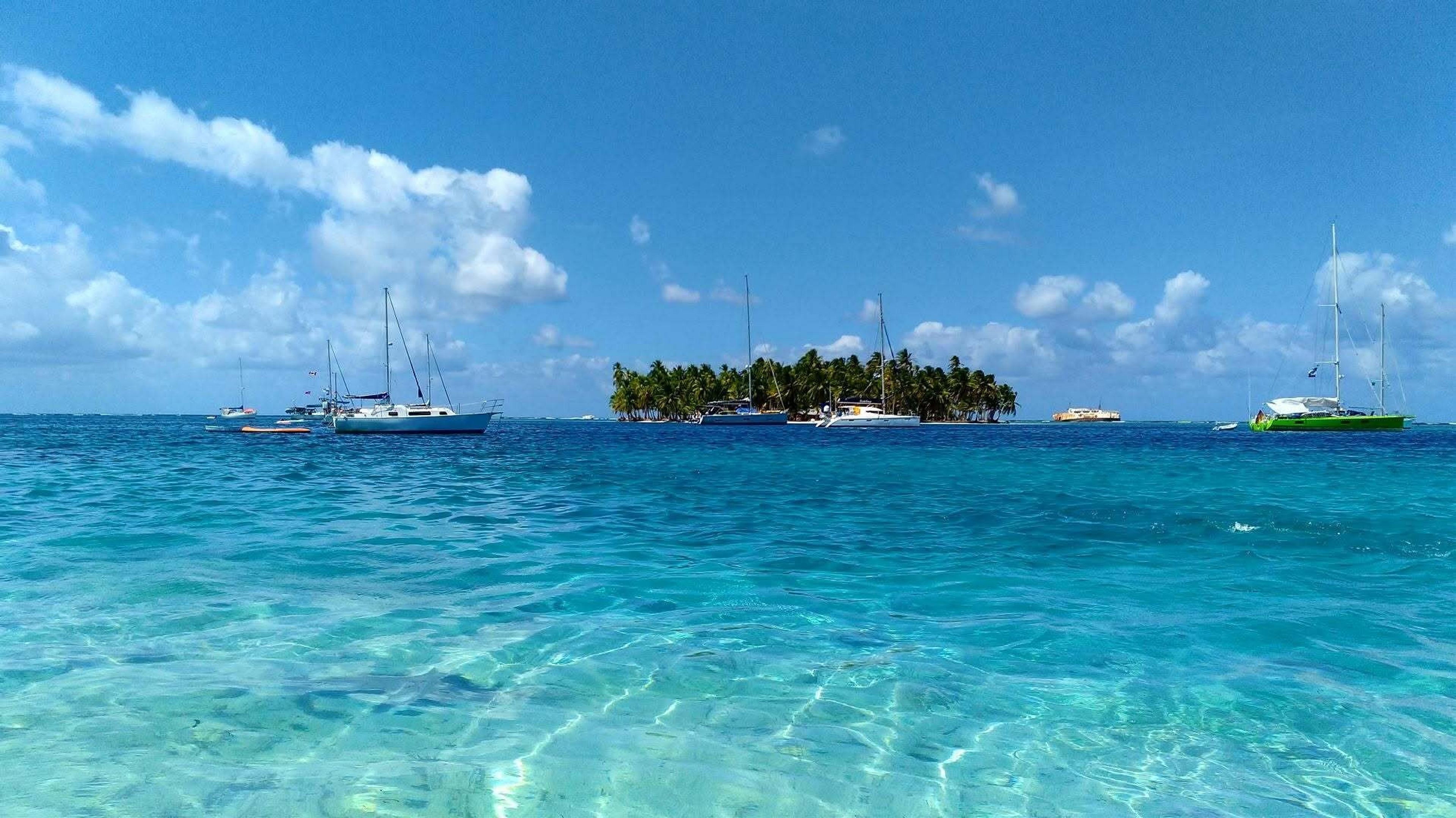 Sailboats float near a small palm-covered island in the clear waters of the San Blas Islands in Panama.