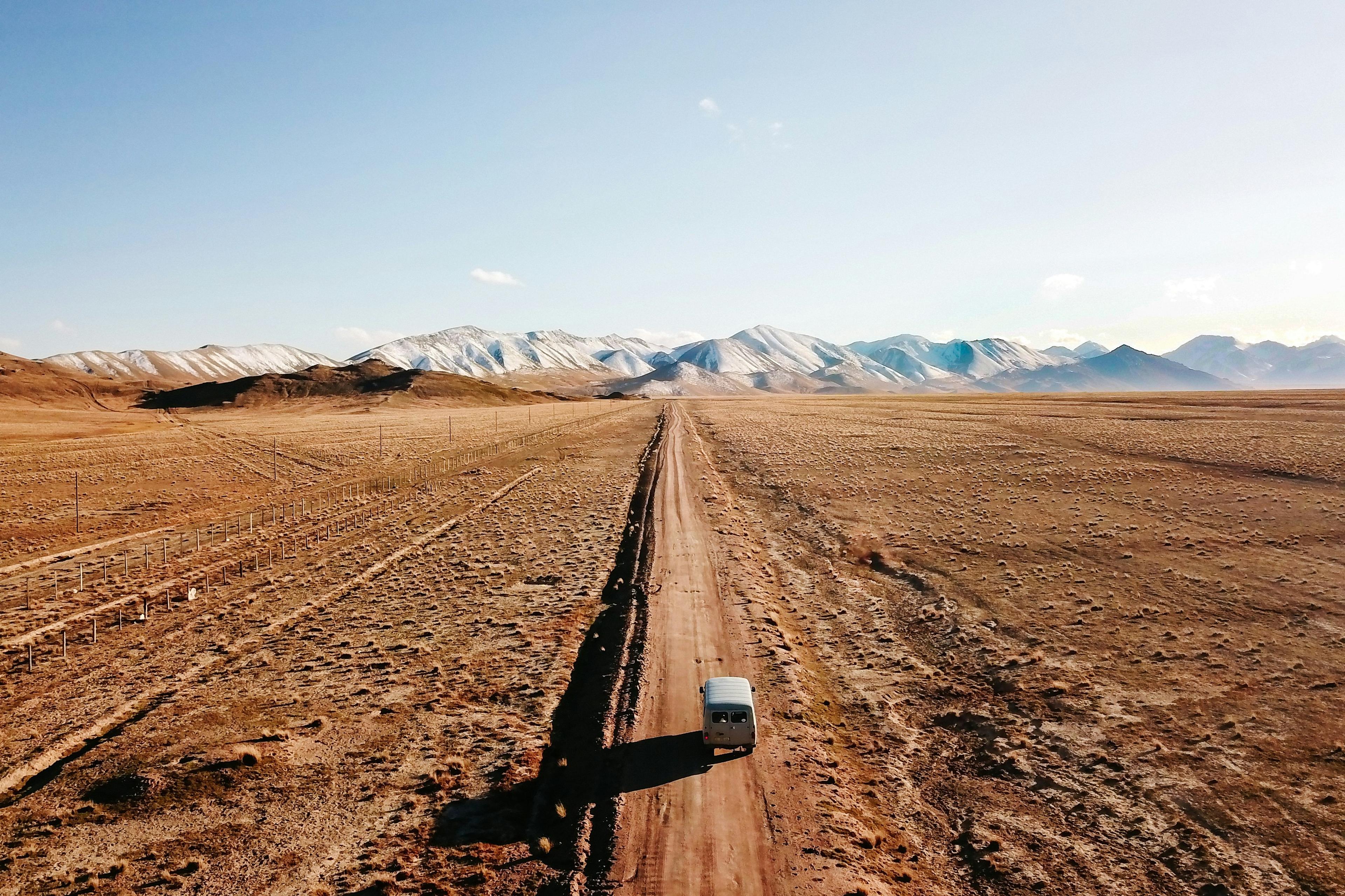 A van travels down a long dirt road in the vast and desolate landscape of Mongolia with snow-capped mountains in the distance.