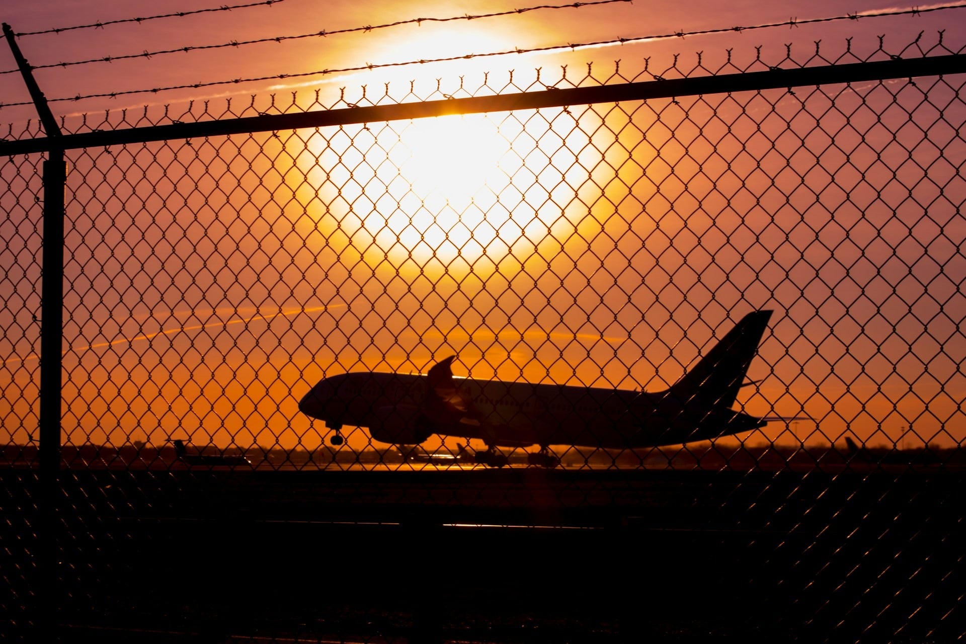 An airplane is silhouetted against a vibrant sunset behind a chain-link fence at an airport runway.