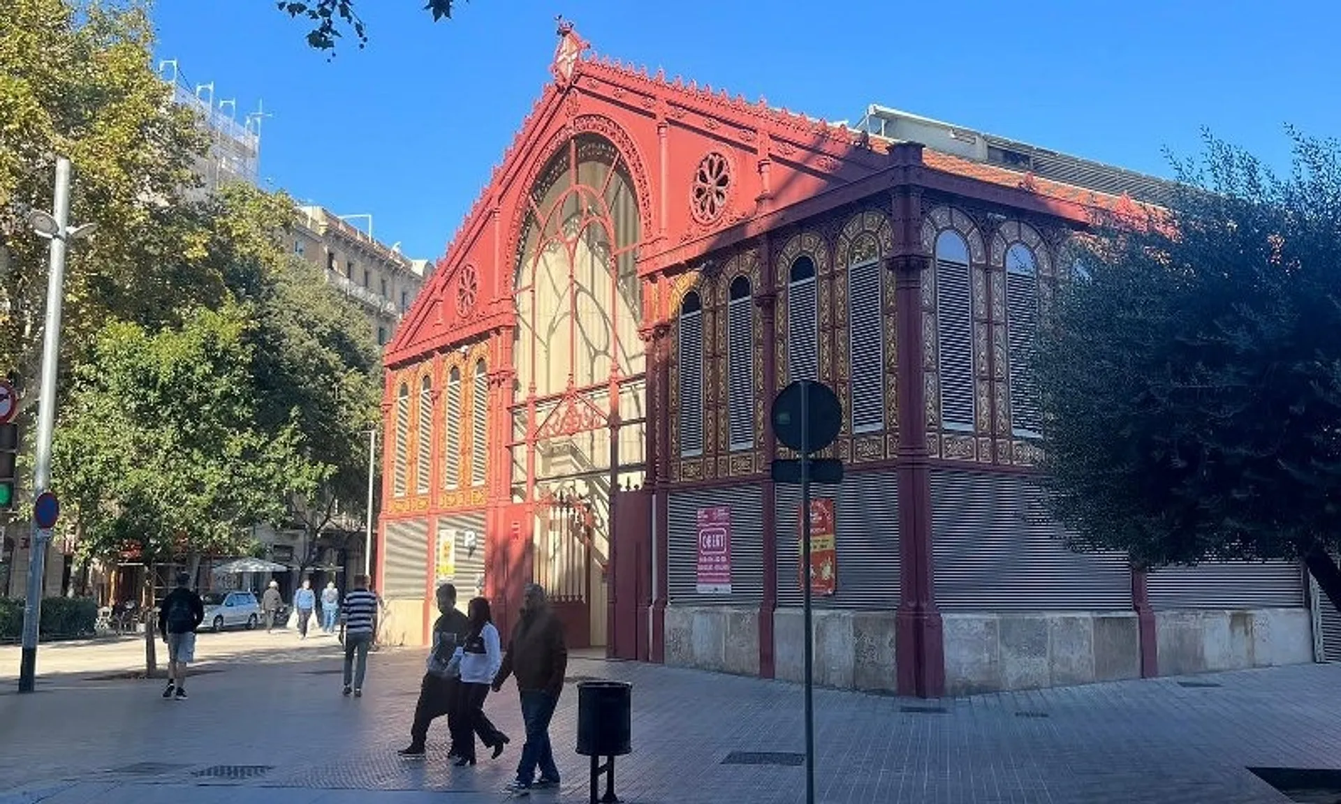 The Mercat de Sant Antoni in Barcelona features a beautifully ornate facade with a mix of iron and glass structures, surrounded by pedestrians on a sunny day.