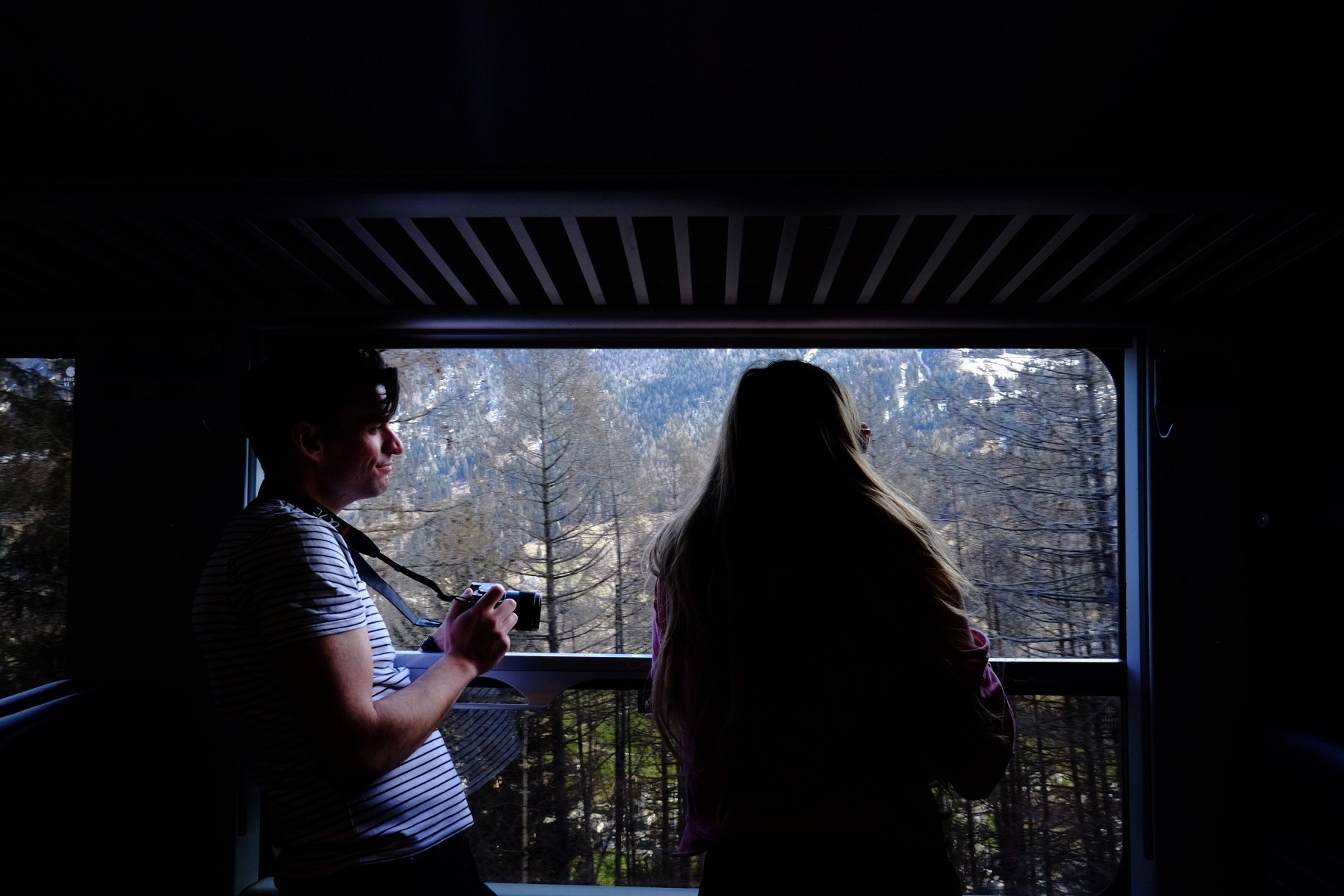 Two people enjoy the scenic mountain view through a train window, possibly somewhere in the Swiss Alps.