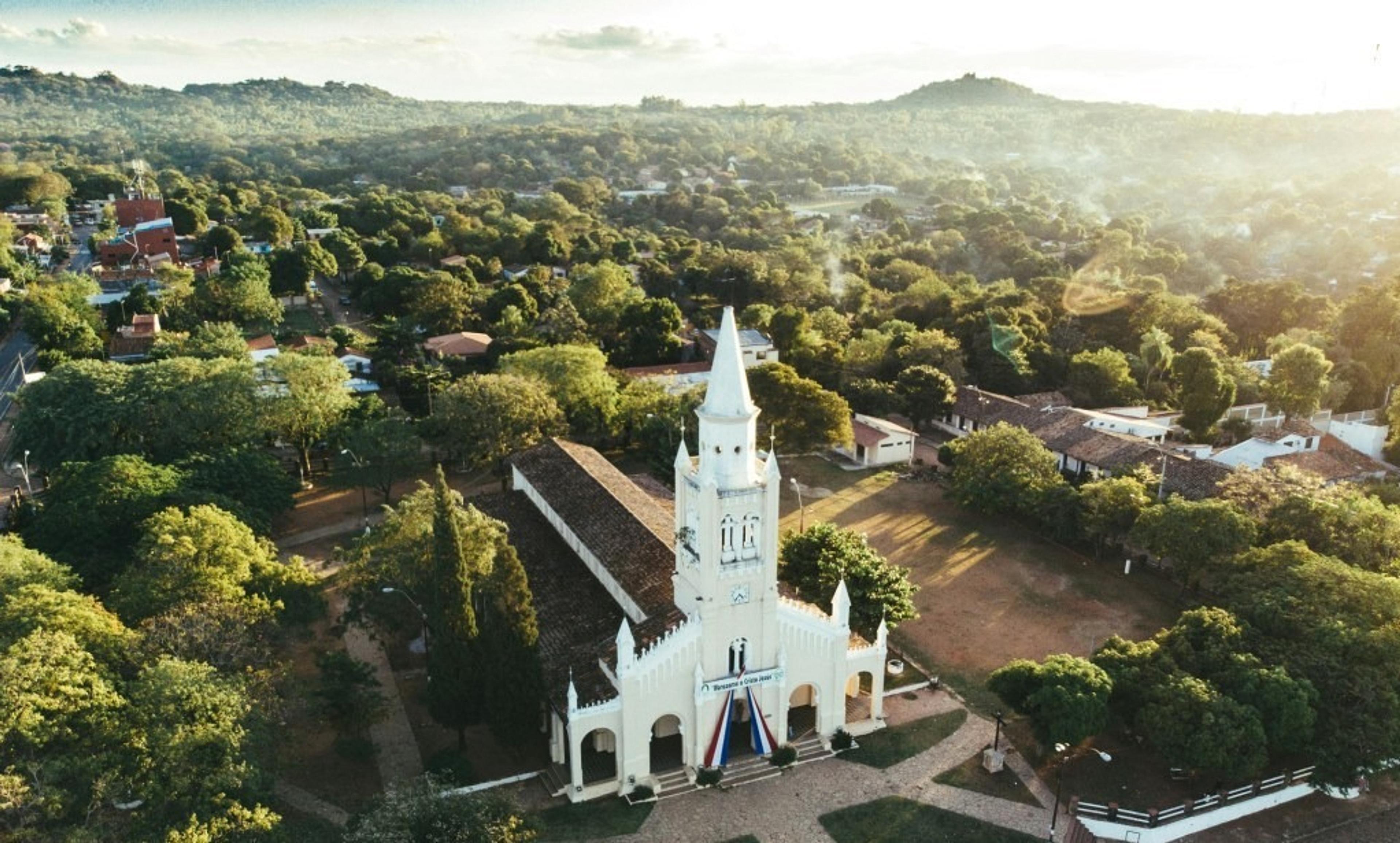 An aerial view of the San Buenaventura Cathedral adorned with a Paraguay flag in Yaguarón, Paraguay, surrounded by lush greenery and distant hills.