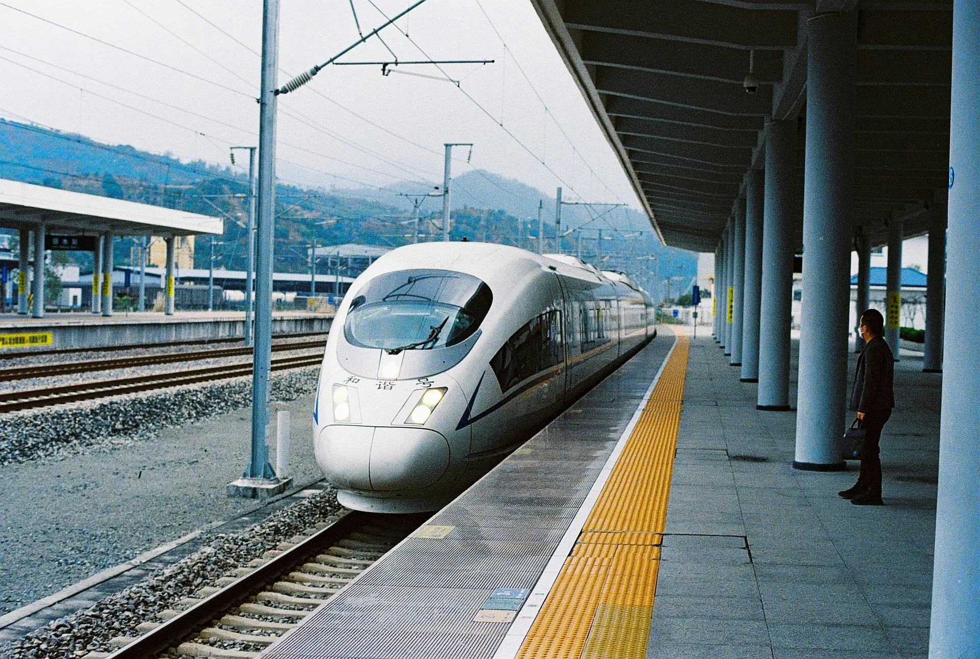 A high-speed train arrives at a platform in a station in China, with mountainous terrain in the background.