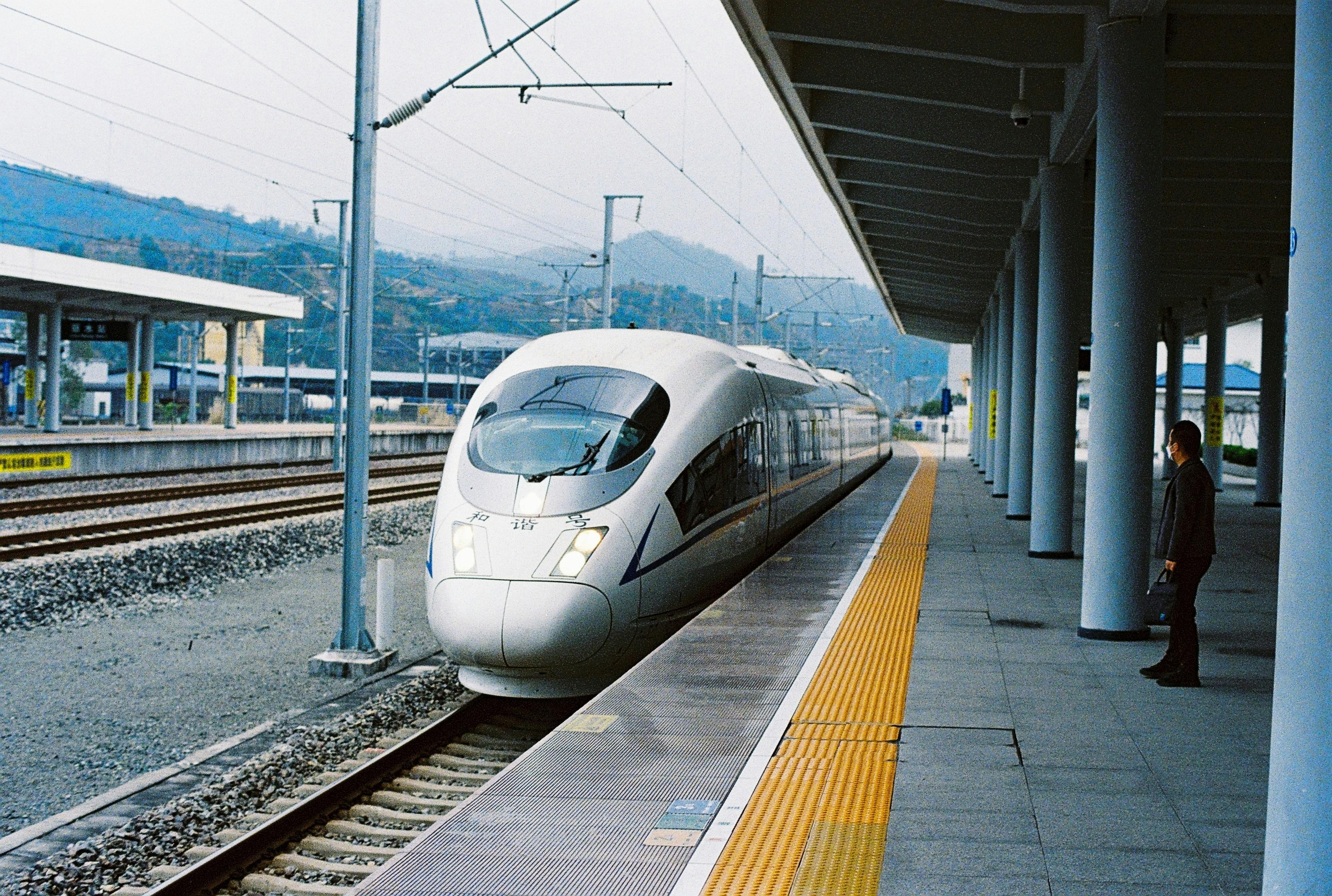 A high-speed train arrives at a platform in a station in China, with mountainous terrain in the background.