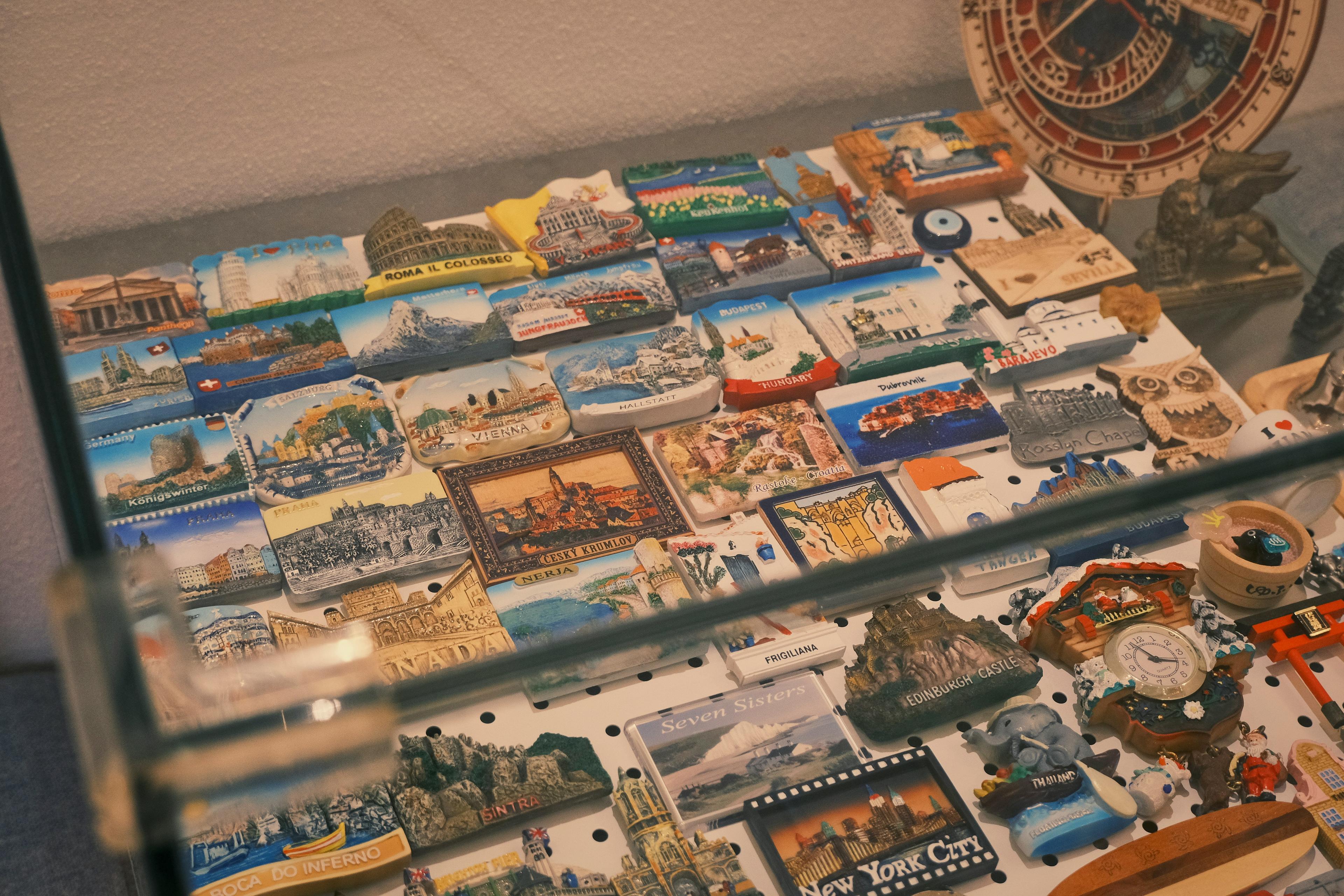 A glass display case contains a collection of colorful souvenir magnets from various worldwide locations such as Rome, Budapest, Vienna, and New York City.