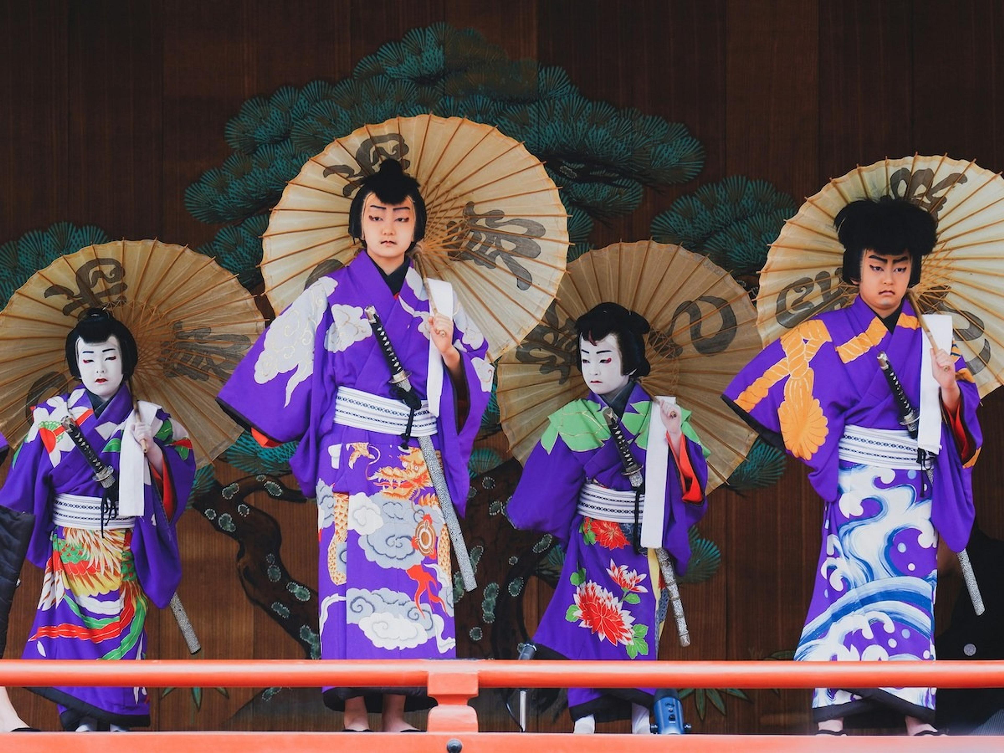 Performers in vibrant traditional attire stand with umbrellas in a Kabuki theater setting in Japan.
