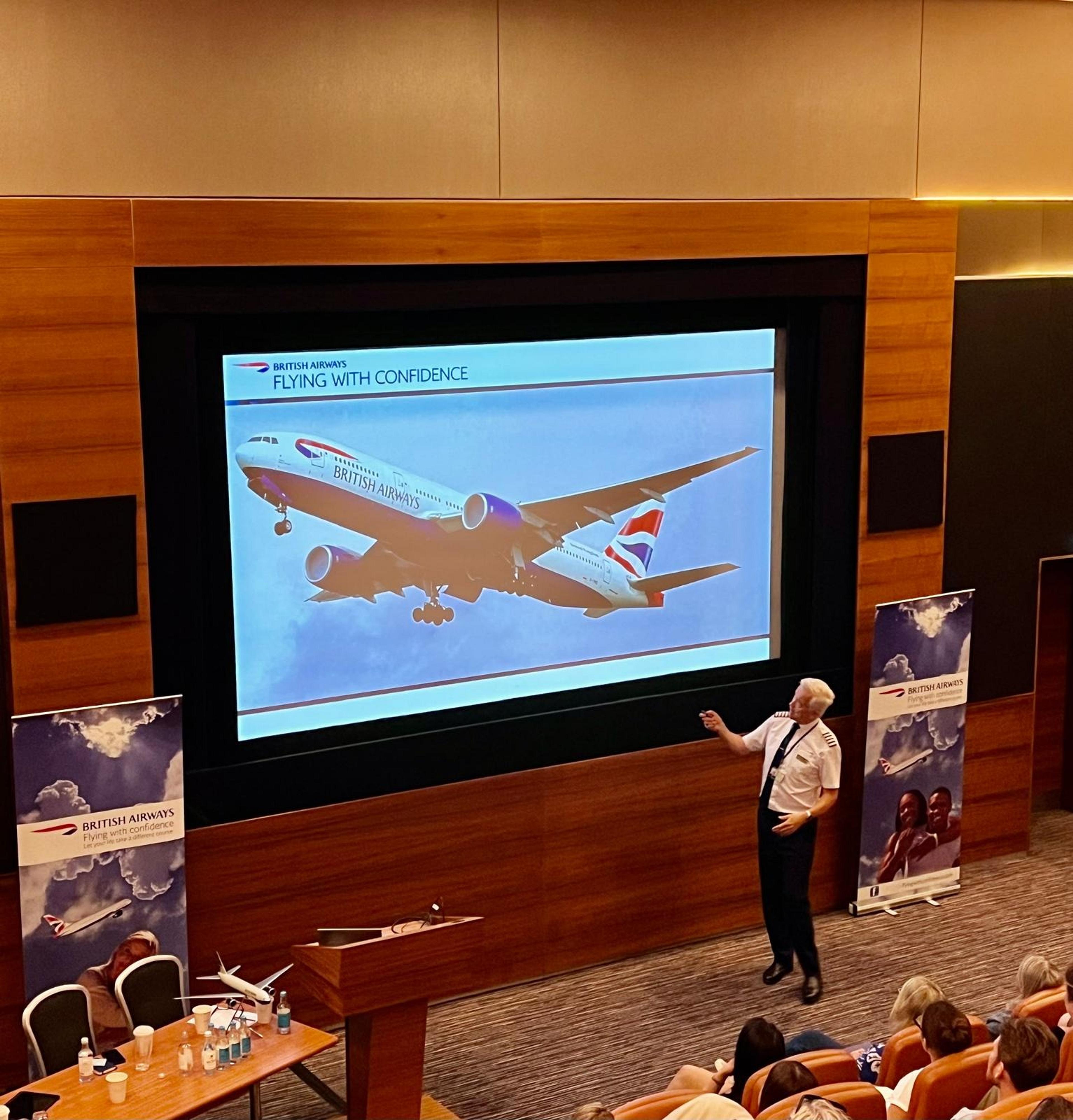 In a lecture hall, a speaker presents a "Flying With Confidence" program image featuring a British Airways plane to an audience.